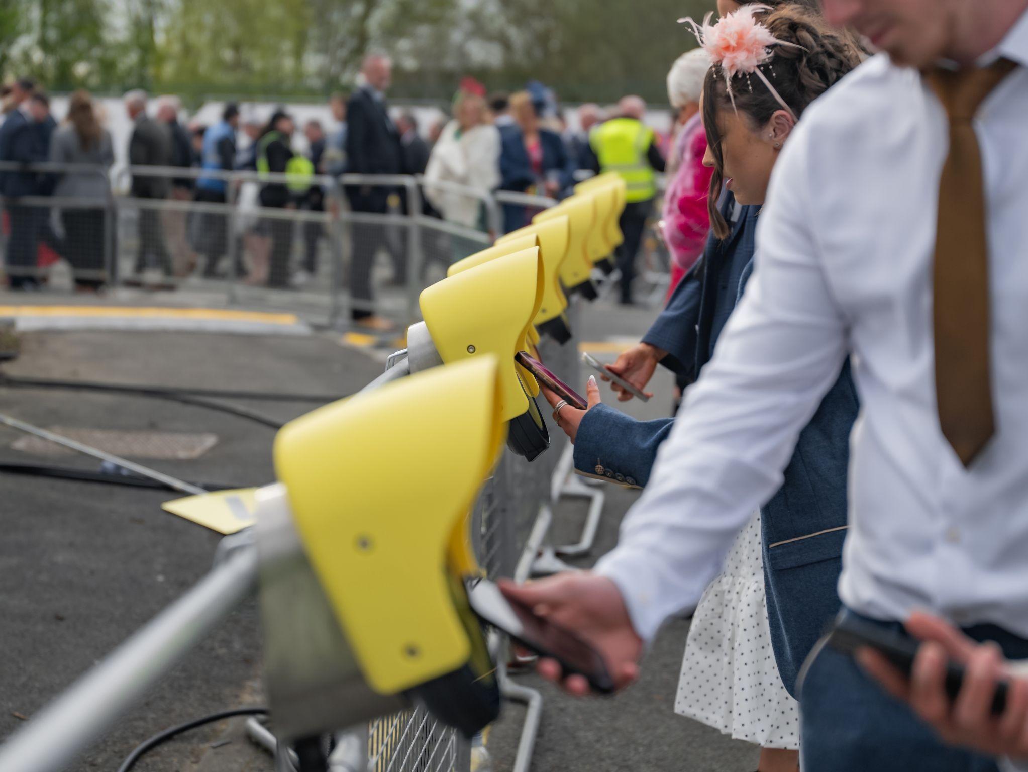 People using yellow ticketing terminals at an outdoor event, with a crowd visible in the background.