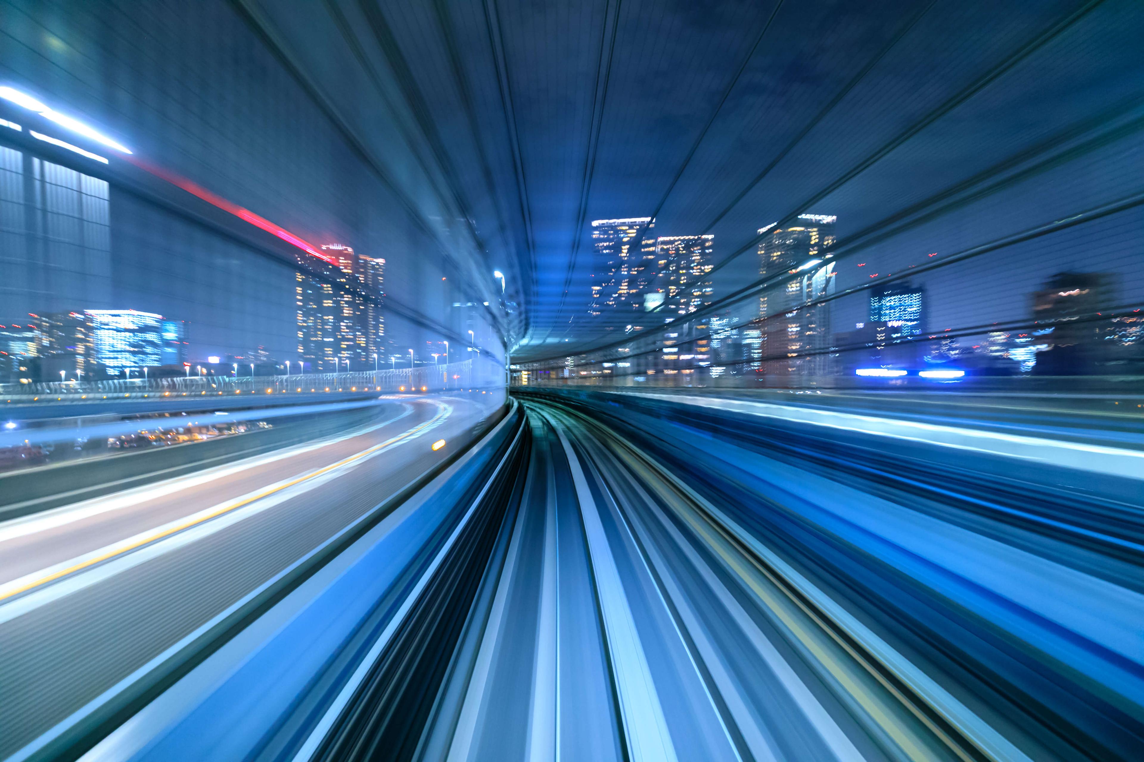 Blurred long-exposure photo of a train speeding through a cityscape at night, with glowing lights and streaks creating dynamic motion.