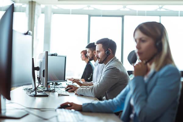 People wearing headsets working at computers in a bright office, focusing on their screens, with large windows in the background.