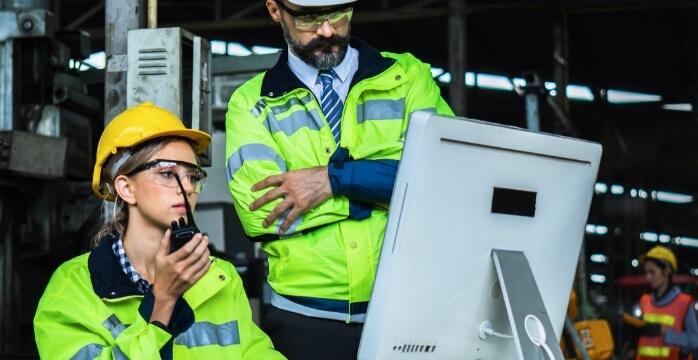 Two workers in safety gear and helmets, one using a computer and holding a walkie-talkie, in an industrial setting.