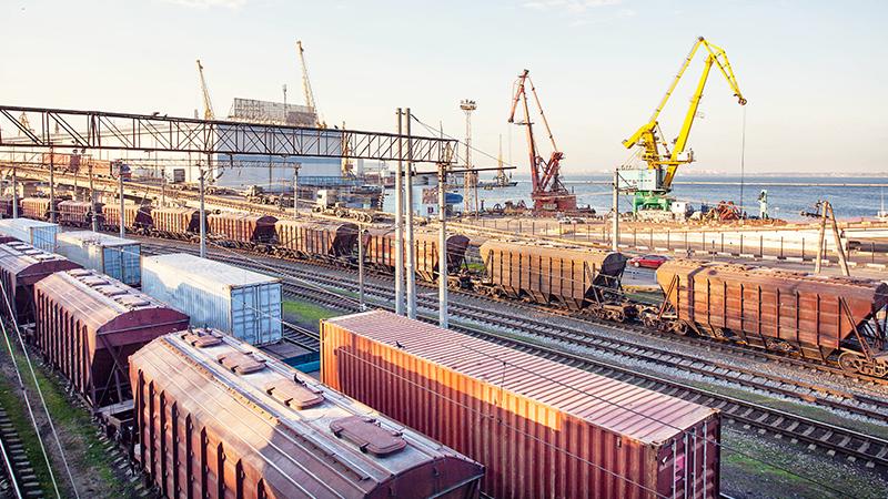 Freight trains on railway tracks near a busy industrial port with cranes and shipping containers by the waterfront.