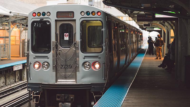 A silver train stopped at an elevated urban station platform, with passengers waiting and a few people visible inside the train.