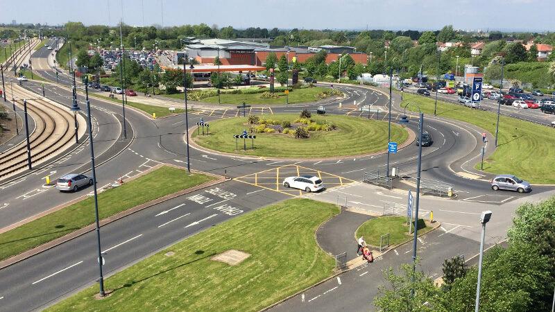 Aerial view of a busy roundabout with multiple lanes, cars, and surrounding greenery on a sunny day.