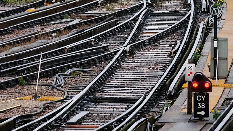 Railway tracks with multiple switches and signals, surrounded by gravel and overgrown grass, indicating a complex rail junction.