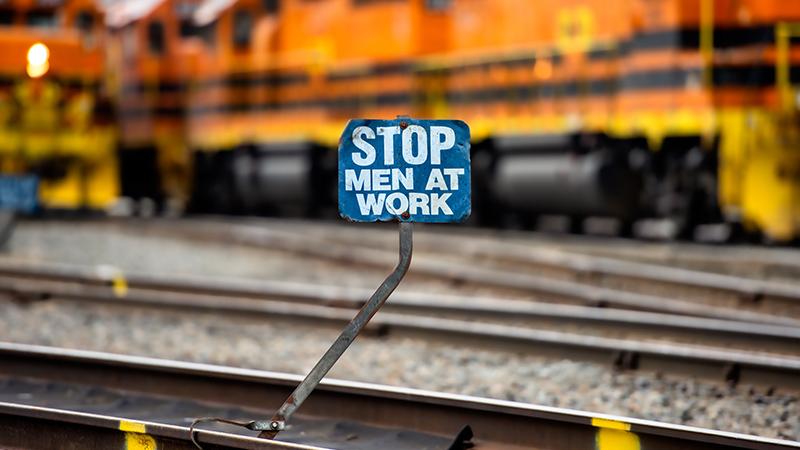 A "STOP MEN AT WORK" sign on a metal pole stands beside railway tracks, with blurred orange trains in the background.