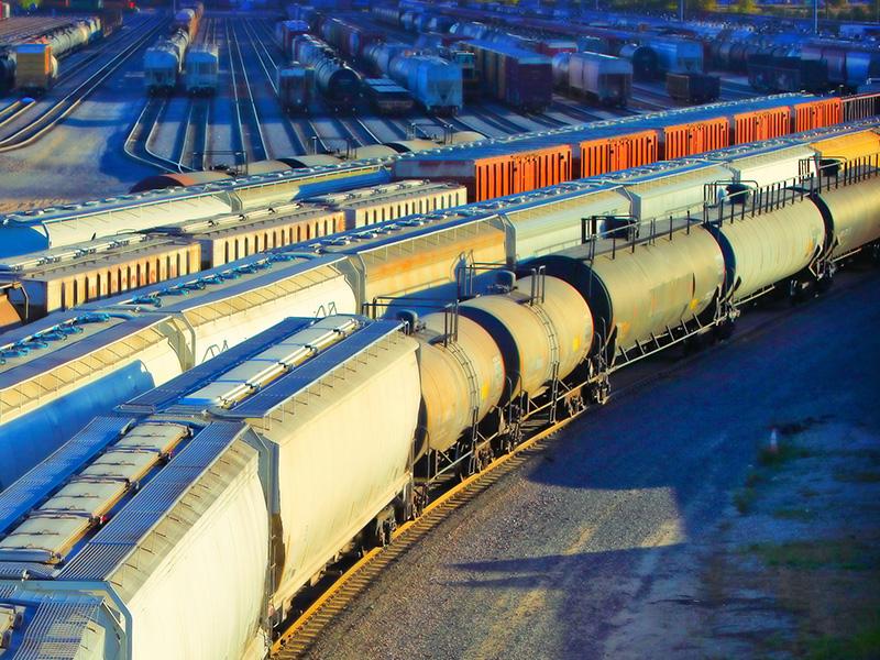Freight train cars and tankers lined up on multiple tracks in a rail yard, with vibrant lighting and shadows.