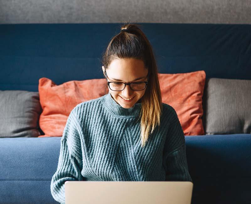 A woman with glasses and a ponytail sits on a sofa, smiling at a laptop. She wears a blue sweater, with cushions in the background.