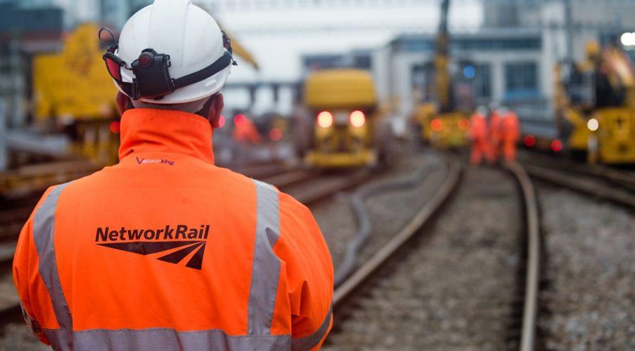 Person in an orange "Network Rail" jacket stands near railway tracks, with workers in the background.