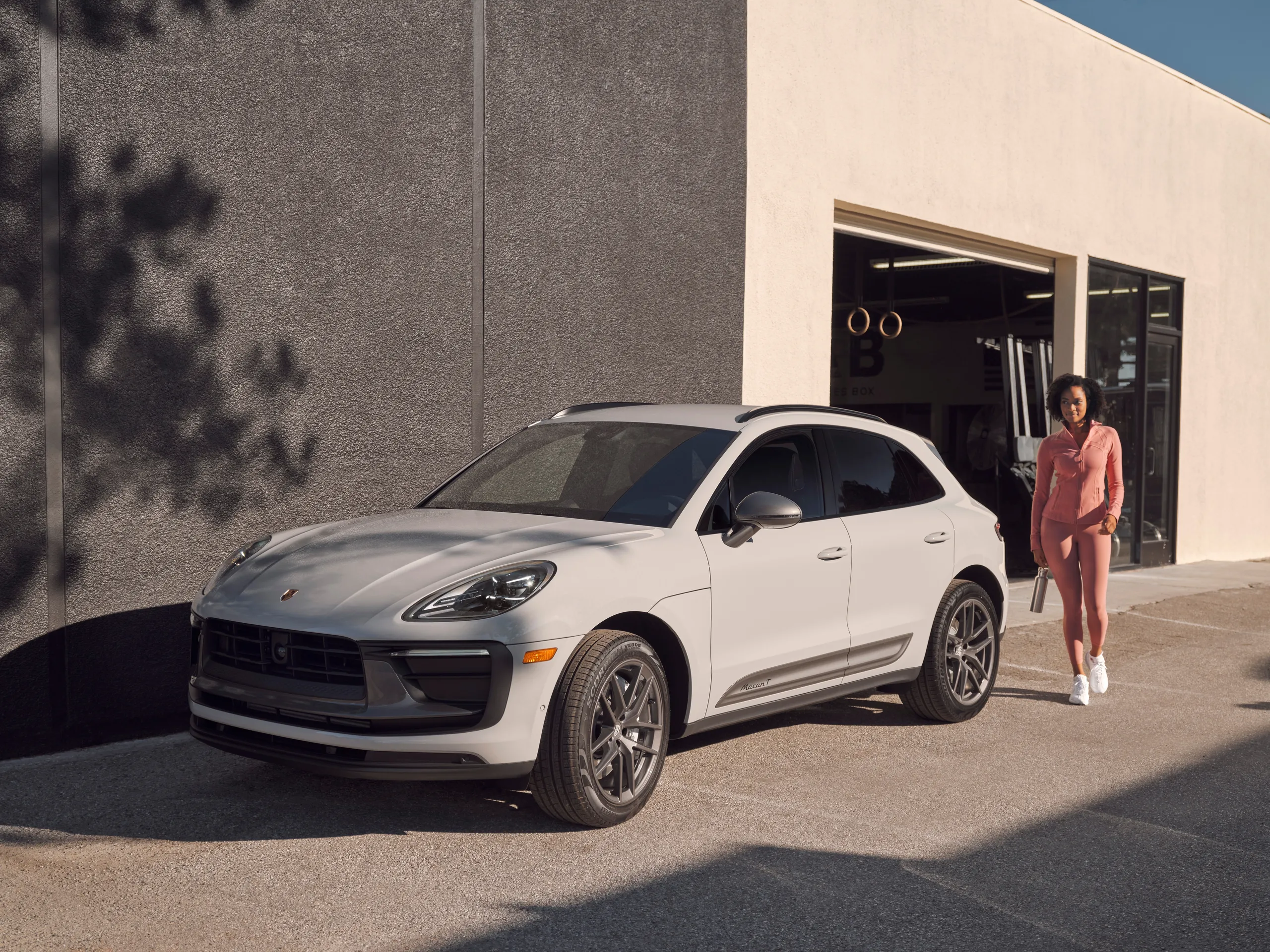 A woman walking up to a grey Porsche Macan parked next to a modern concrete building. 