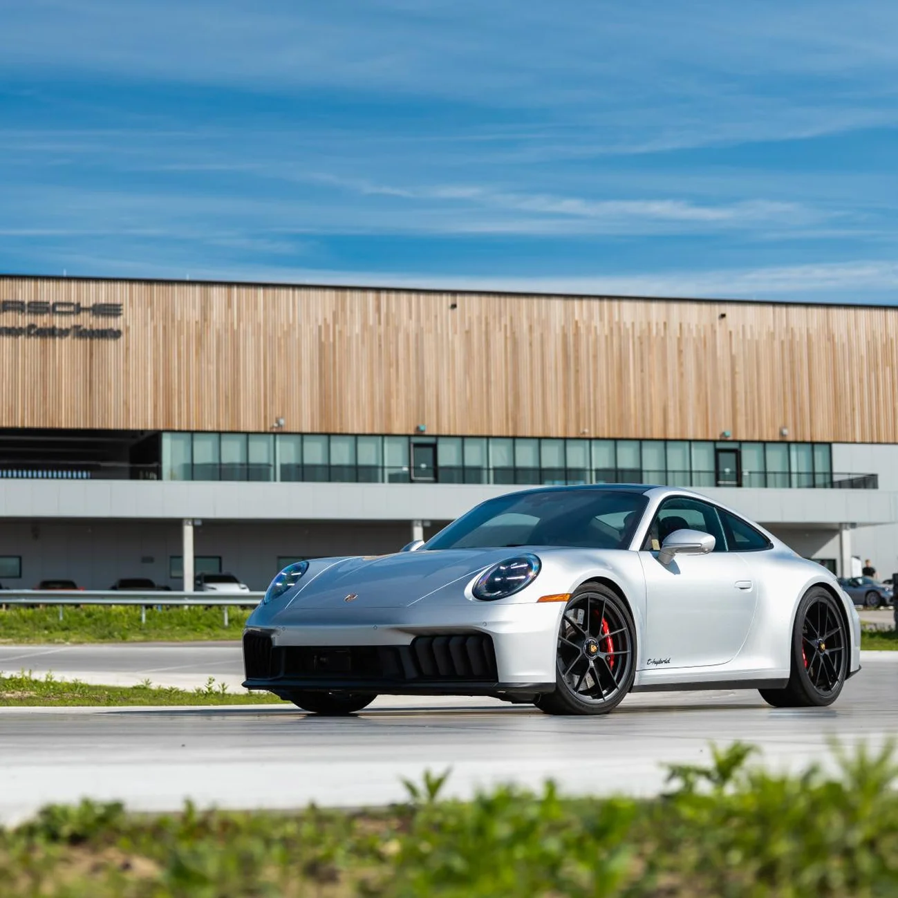 A silver Porsche 911 driving on a circuit with a modern building with a wood facade in the background.