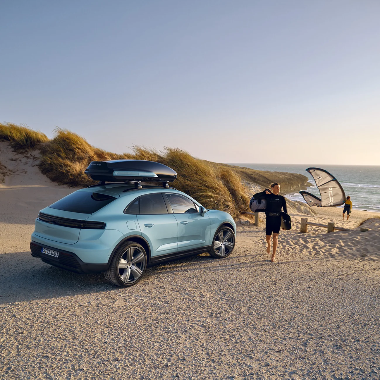 Un hombre camina junto a un Porsche Macan 4 con una caja portaequipajes negra en el techo, aparcado en la playa.