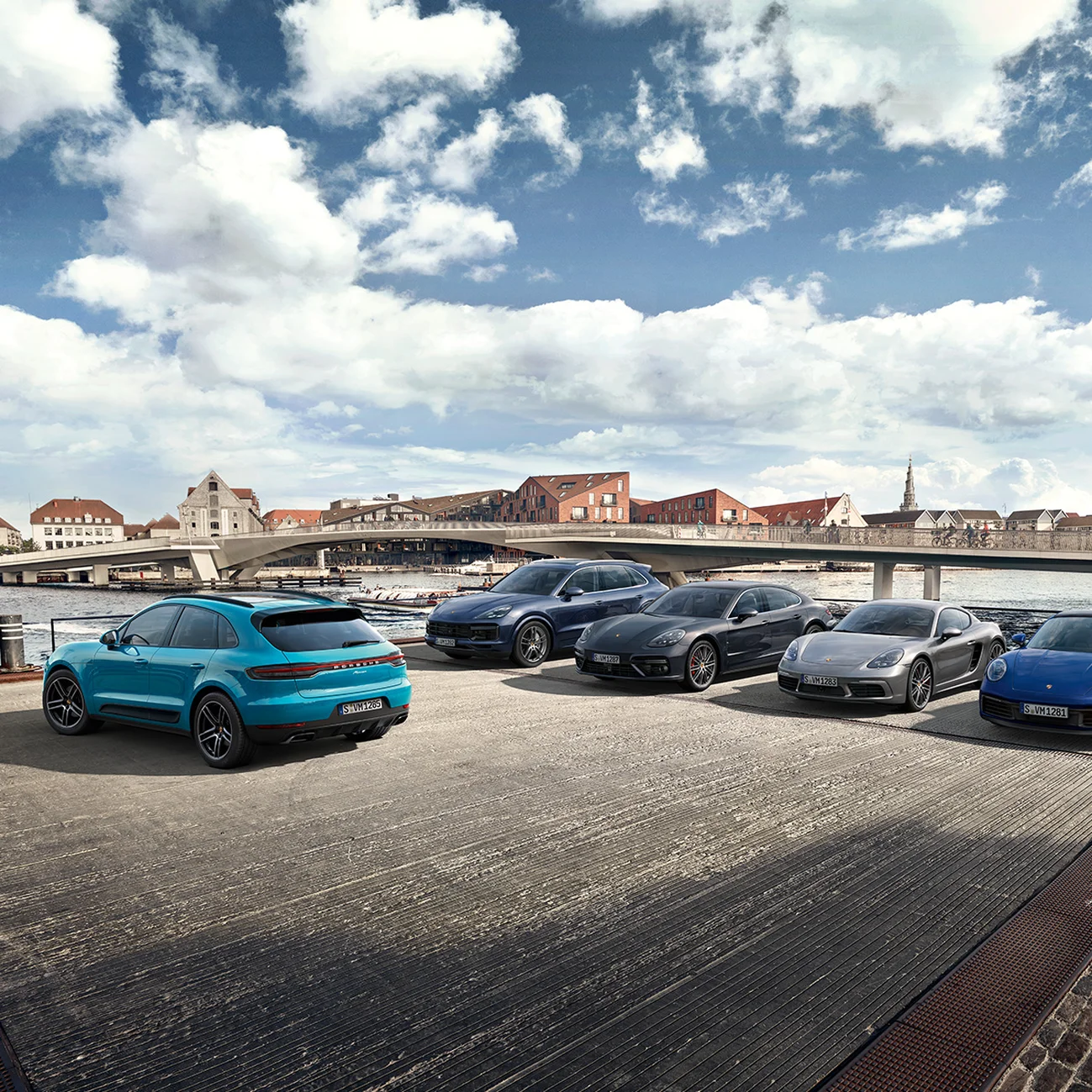Um conjunto de veículos Porsche estacionados perto de um rio e com uma ponte ao fundo.