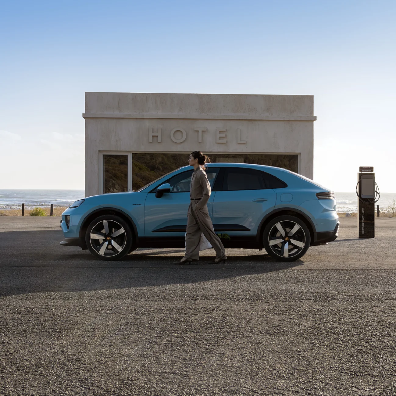 Side shot of a Frozen Blue Metallic Macan Electric parking in front of a modern building by the beach with a woman standing in front of the car.