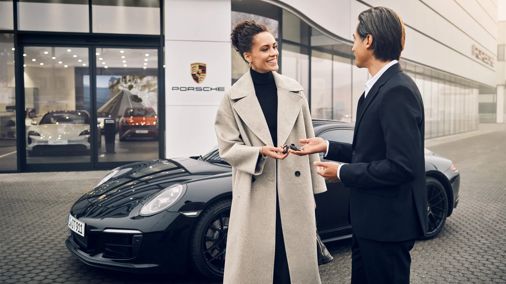 Una mujer recibiendo una llave de coche de un hombre frente a un coche Porsche y un centro Porsche.