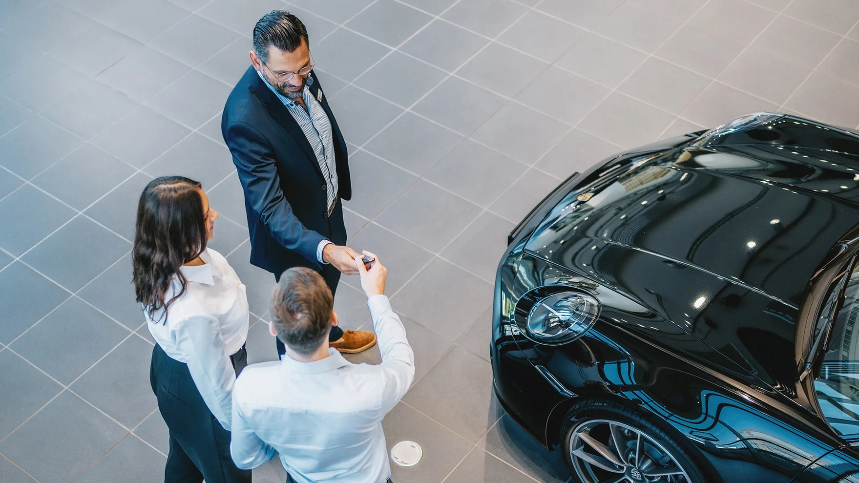 Un homme souriant remet une clé de Porsche à un couple, célébrant la livraison de leur nouvelle voiture dans le cadre du programme Porsche Approved.