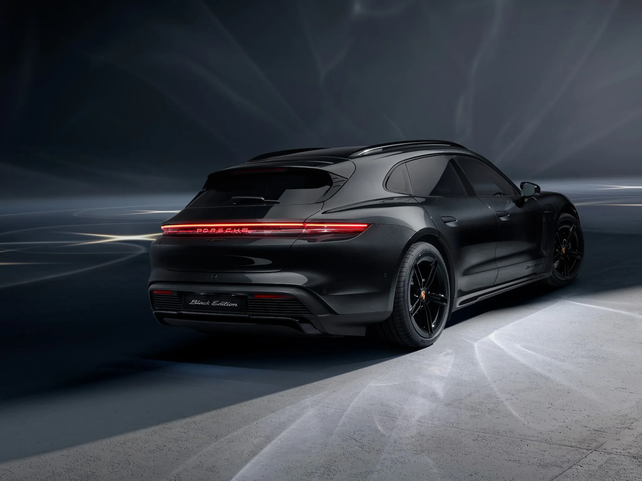A black Porsche Taycan Sport Turismo Black Edition photographed from a rear three quarter angle in a dark studio, featuring illuminated Porsche rear light bar, Black Edition branding, and black performance wheels.