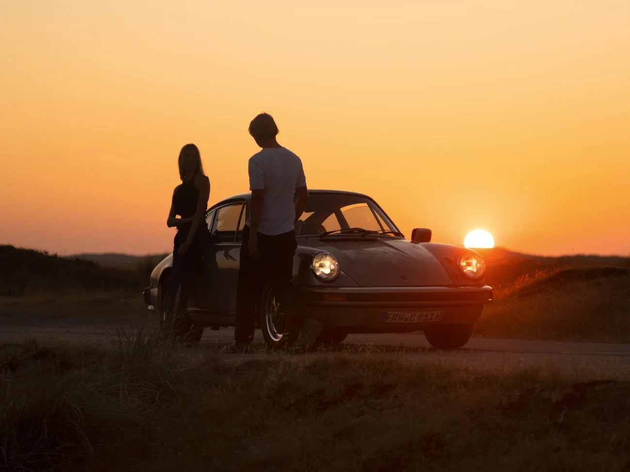A woman and a man stand in front of an old Porsche, with the sunset casting a stunning backdrop.