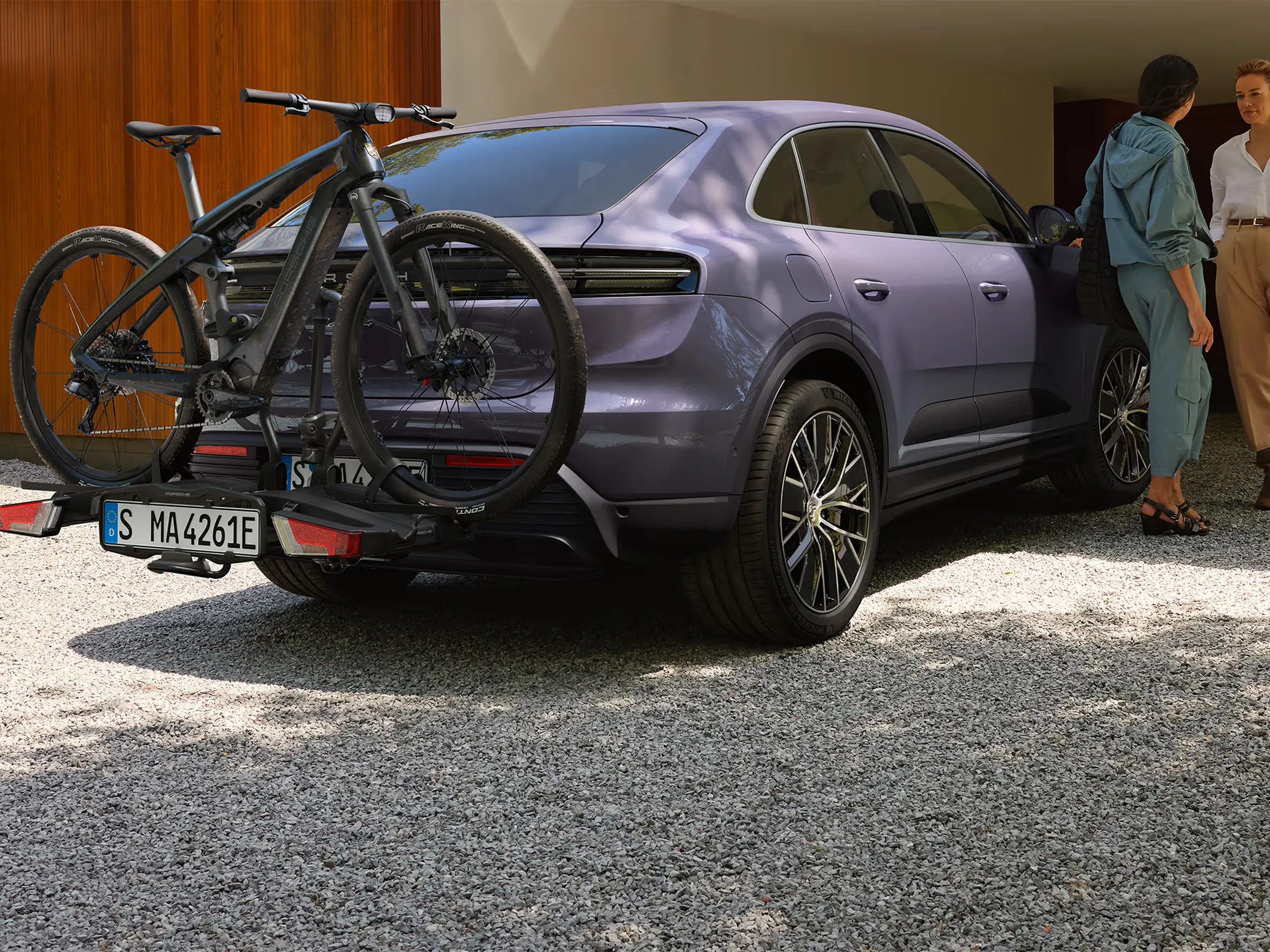 Purple Porsche SUV with rear bike rack and black e-bike mounted on the back, parked in front of a modern garage surrounded by greenery.
