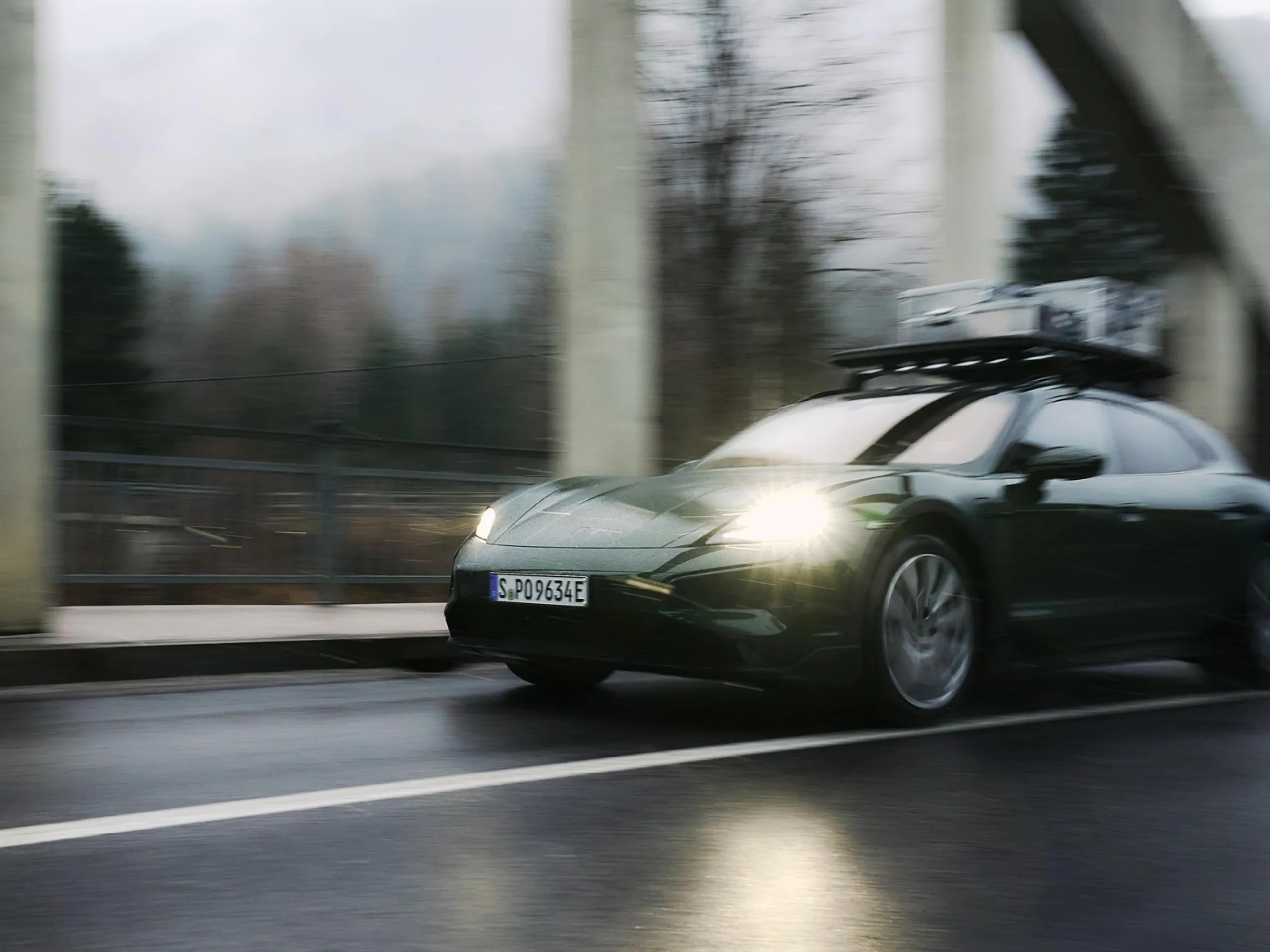 Une Porsche Taycan vert foncé avec un porte-bagages de toit roulant dans des conditions froides et humides.