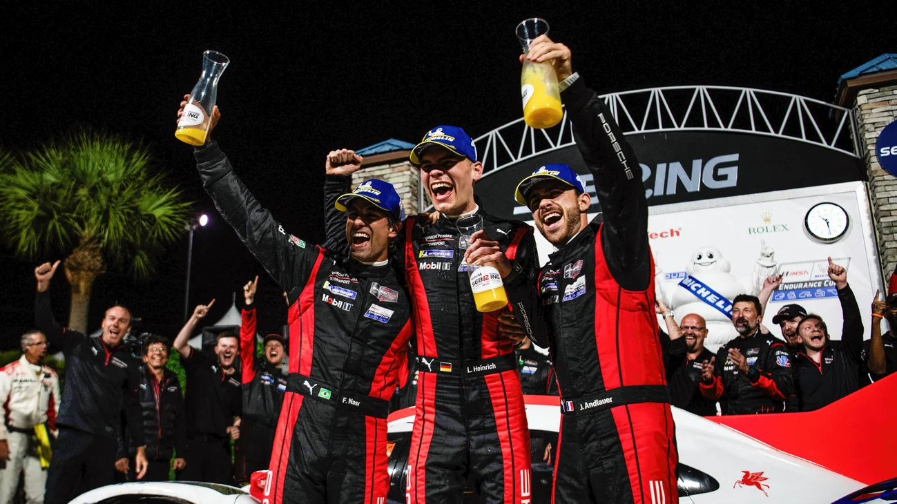 Three race drivers in red and black suits celebrate on a podium, raising bottles as a crowd cheers behind them at night.