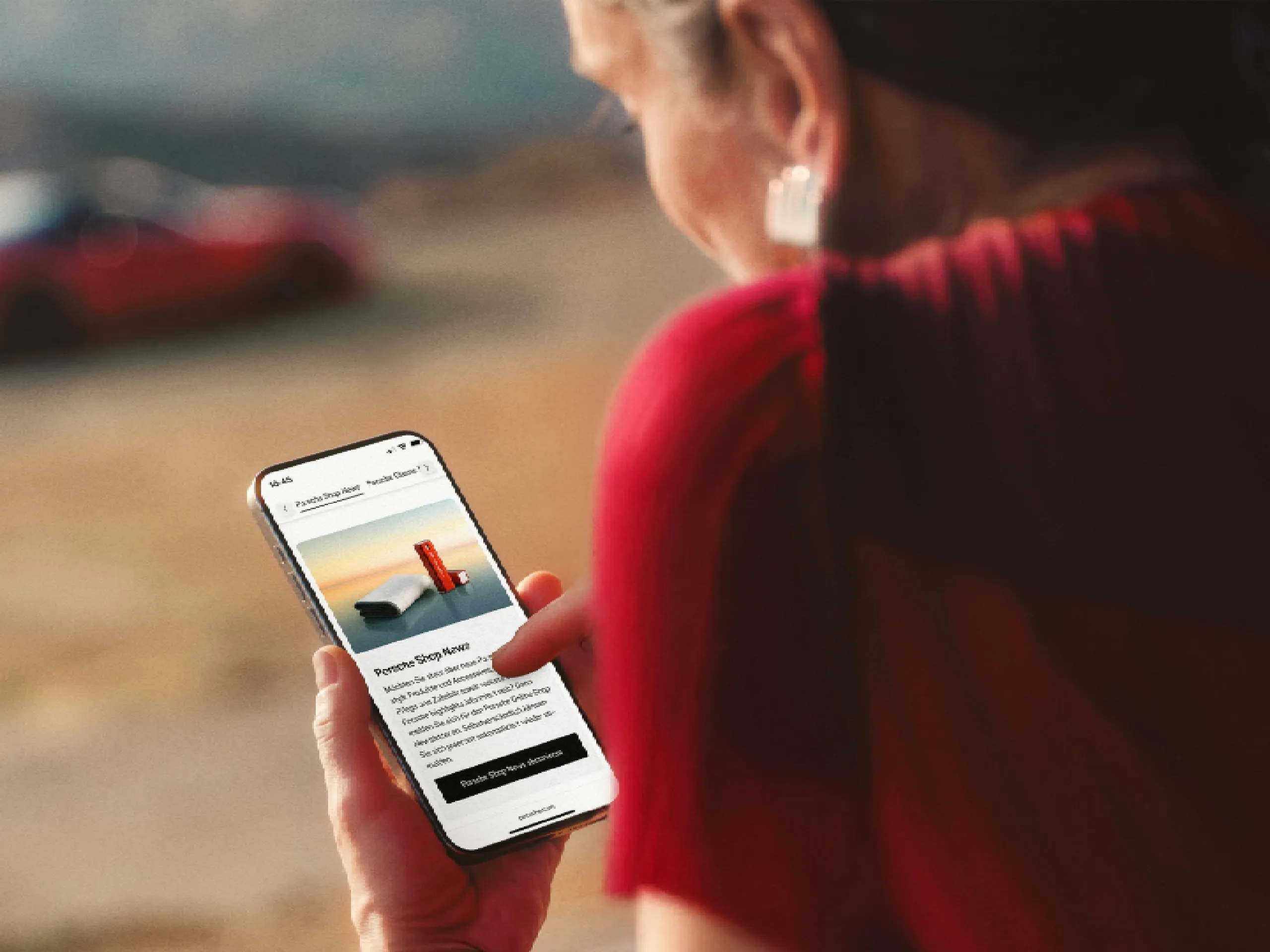 Woman reading the Porsche Shop newsletter on her smartphone; a red Porsche is blurred in the background on a scenic road.