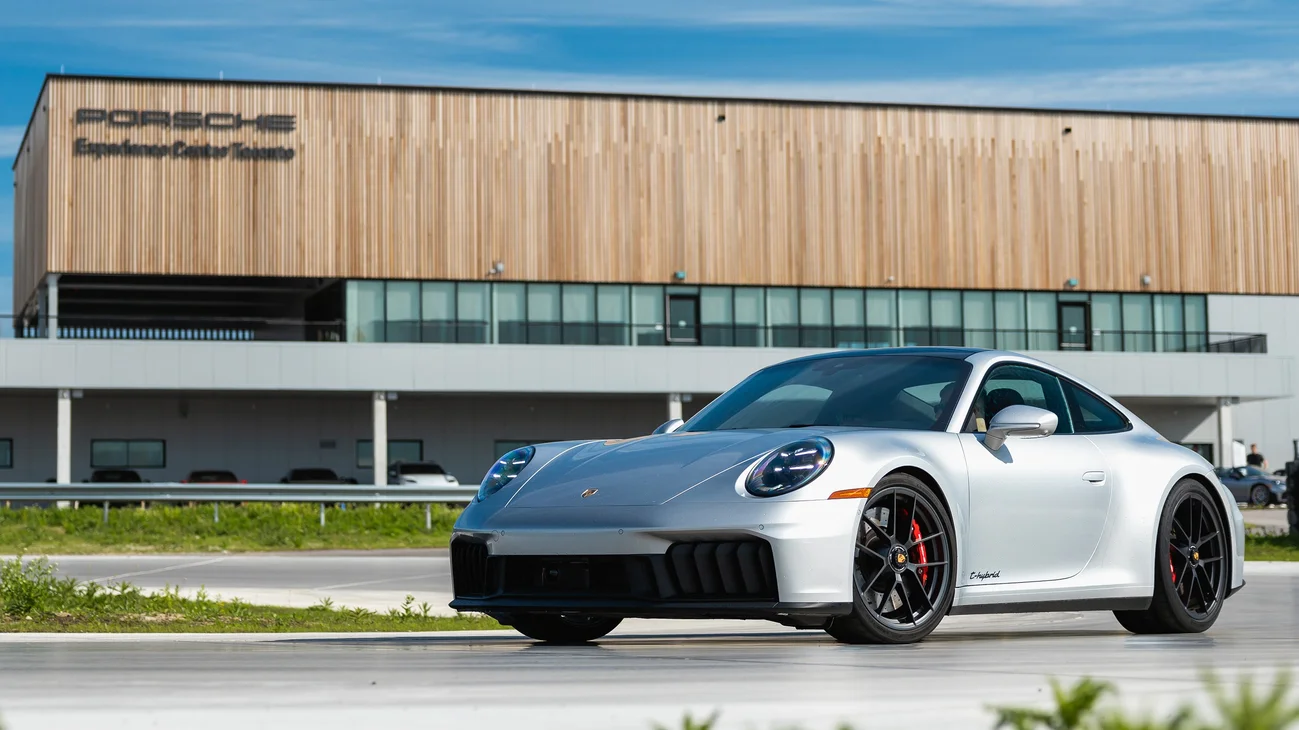 A silver Porsche 911 on the circuit with a modern building with a wood facade in the background.
