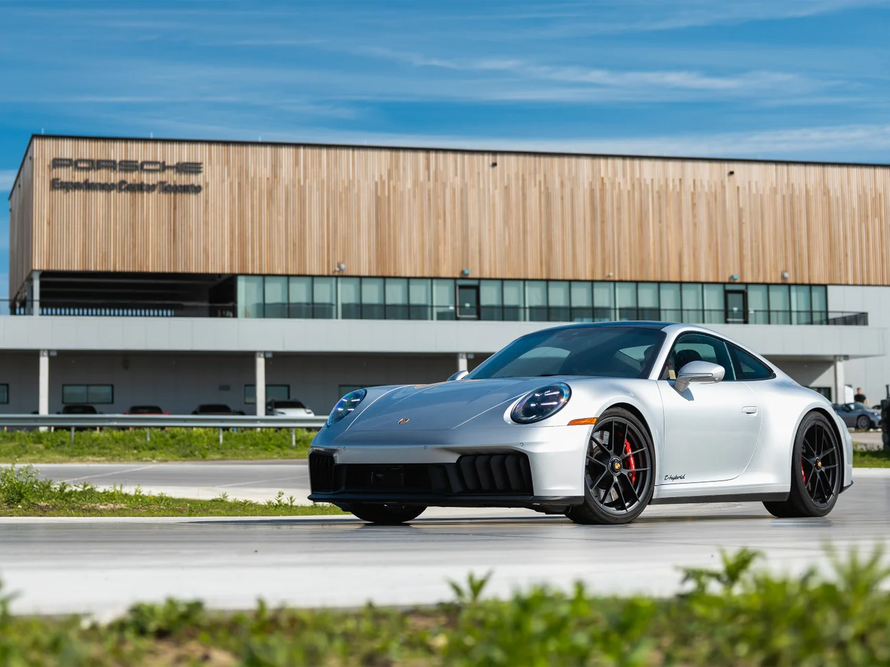 A silver Porsche 911 on the circuit with a modern building with a wood facade in the background.