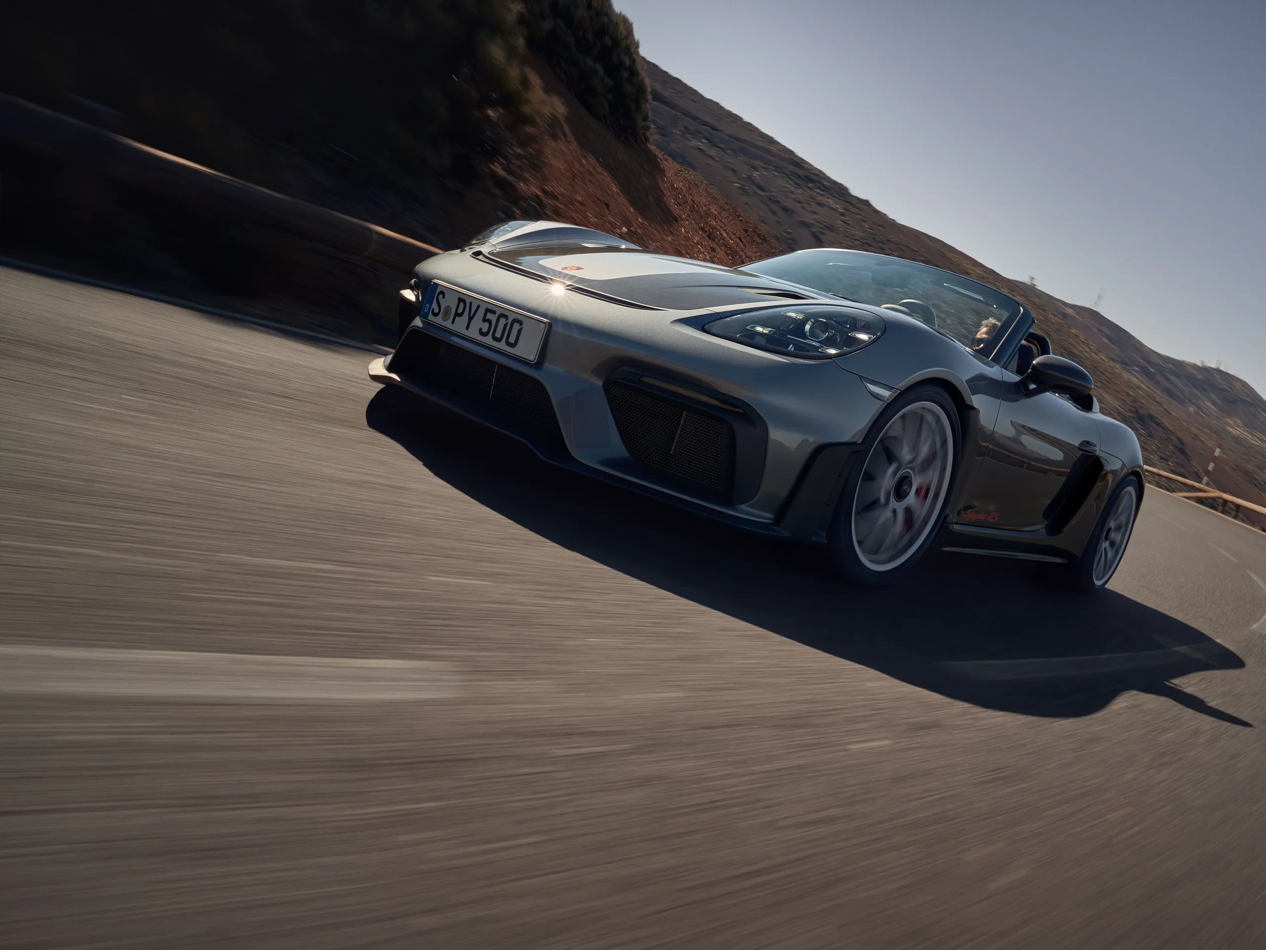 Front shot of a silver 718 Spyder RS driving on a road through a barren desert landscape on a sunny day and going straight around a corner
