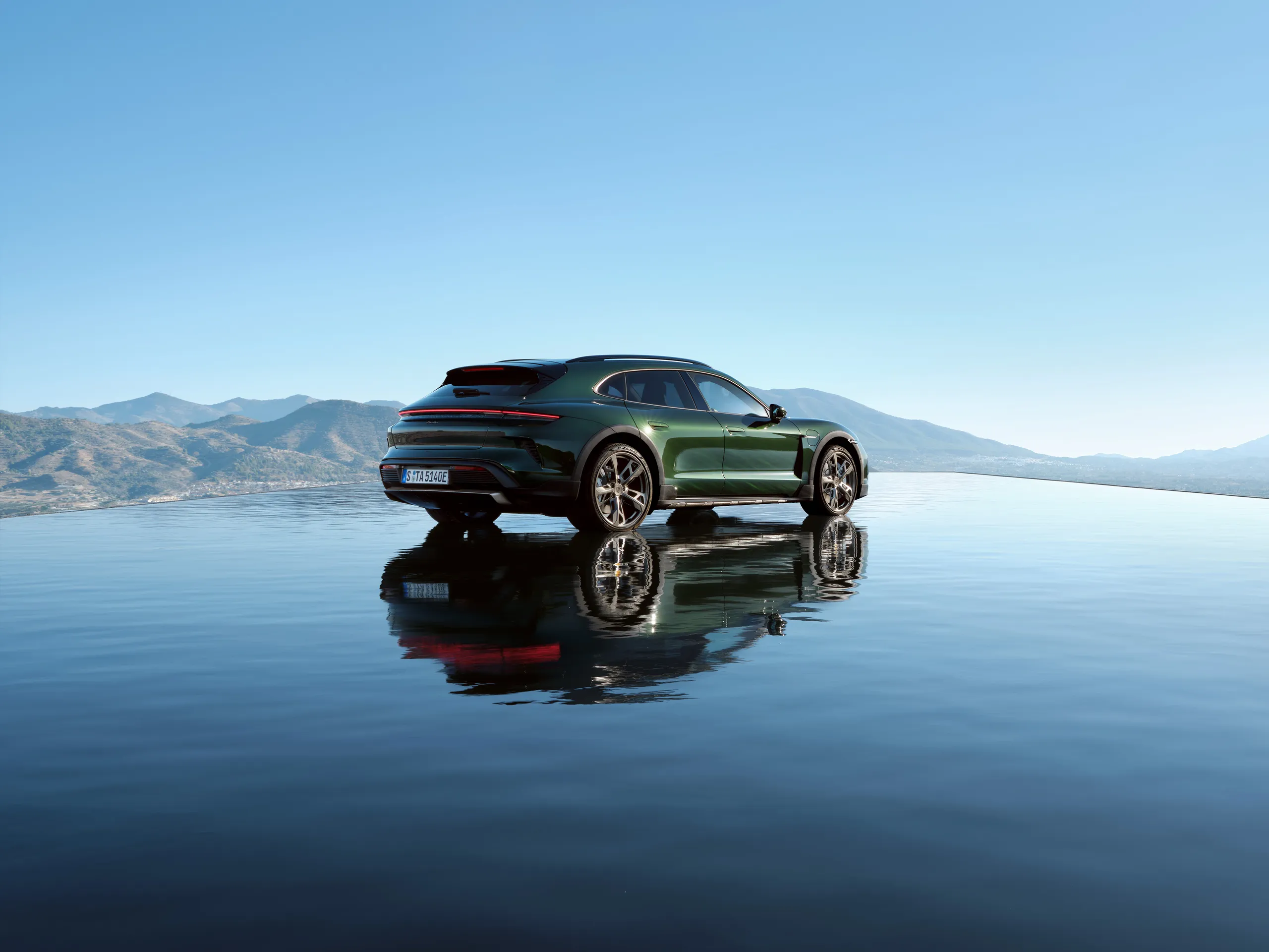 An oakgreenmetallic neo colored Taycan Turbo Cross Turismo stands on a reflective surface in front of a mountain range.