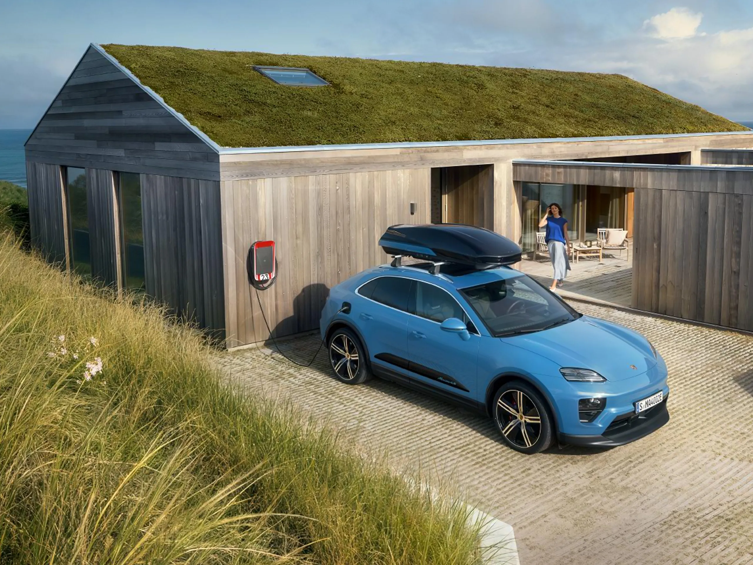 Modern wooden bungalow by the sea, a blue Macan Electric with a roof box parked in front, while a person stands on the terrace in the background.