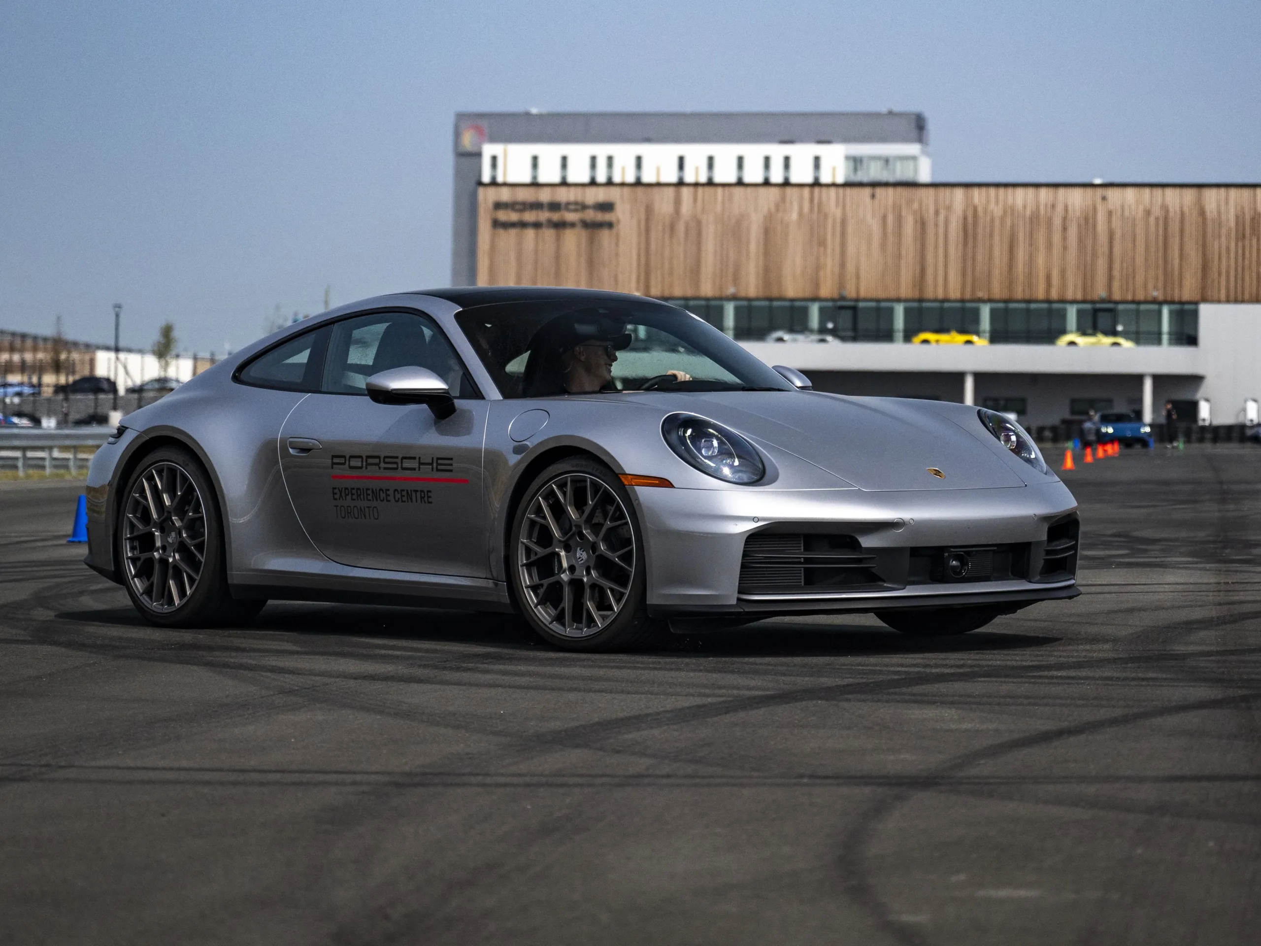Une Porsche 911 argentée roulant sur un circuit, avec en arrière-plan un bâtiment moderne à la façade en bois.