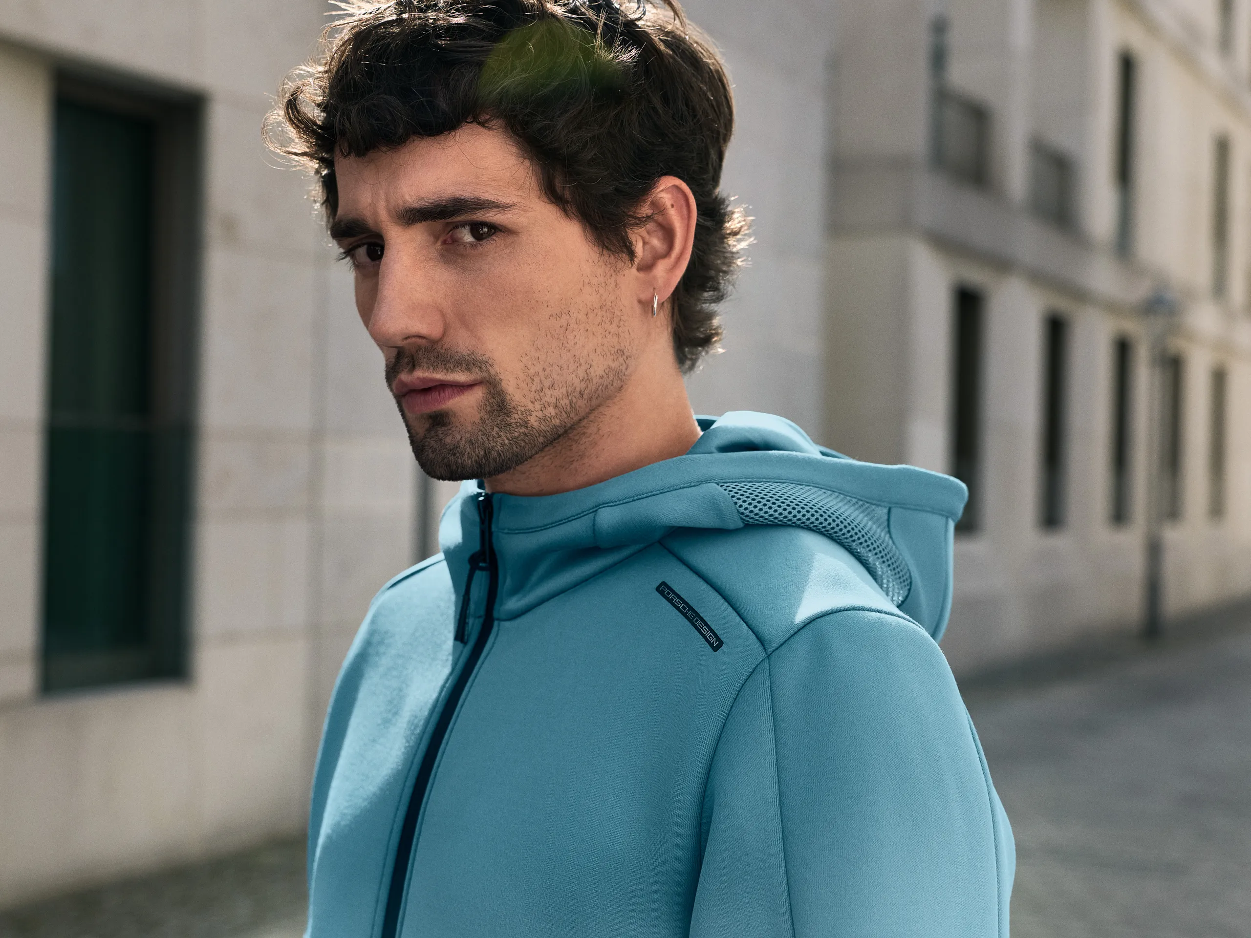 Man wearing a blue Porsche Design sports jacket standing in an urban setting, looking at the camera.