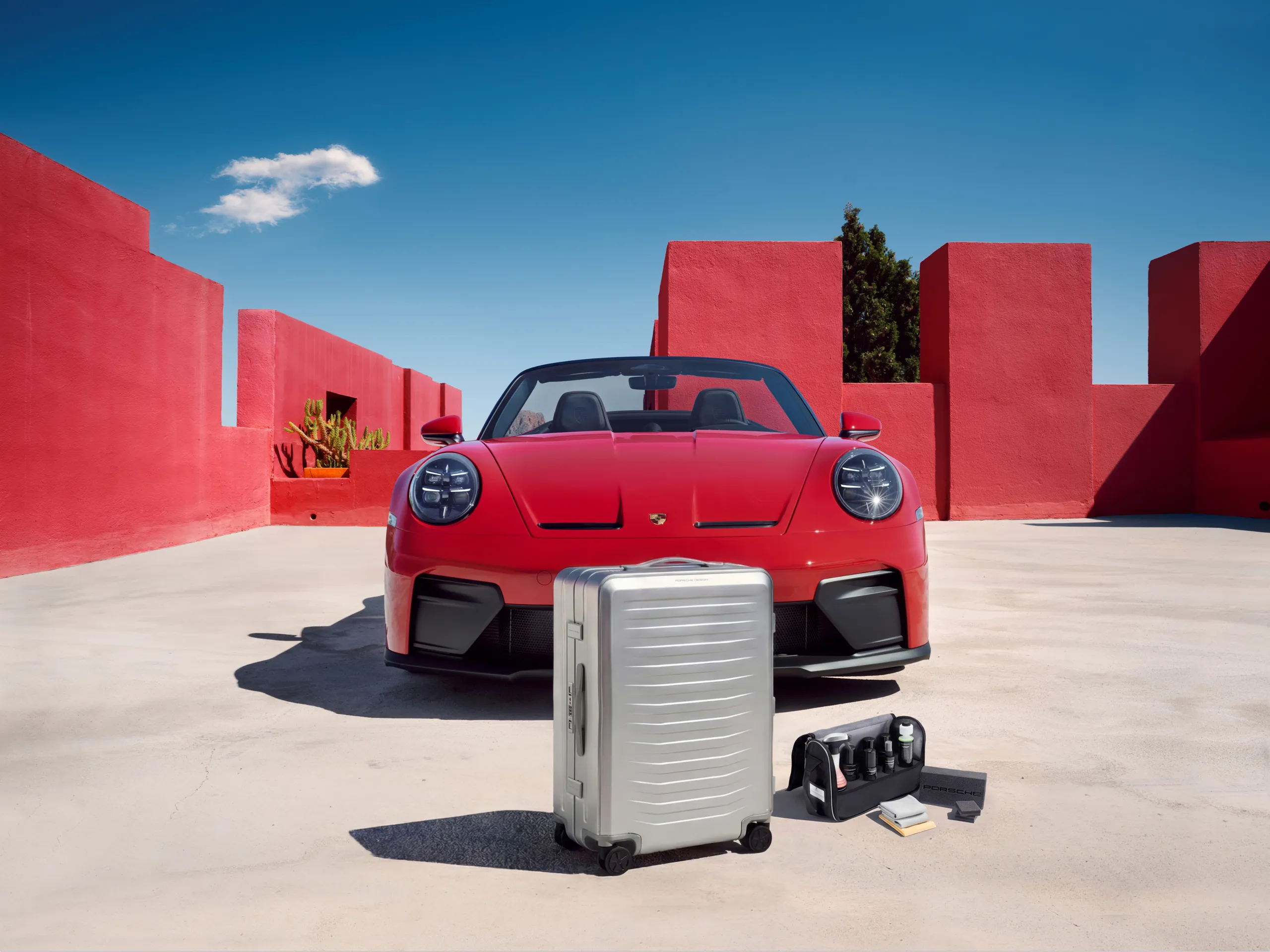 A red Porsche 911 GT3 S/C in front of a red building, with a silver suitcase and a care bag in the foreground.