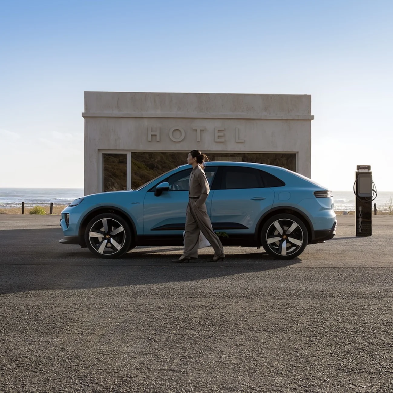 Side shot of a Frozen Blue Metallic Macan Electric parking in front of a modern building by the beach with a woman standing in front of the car.