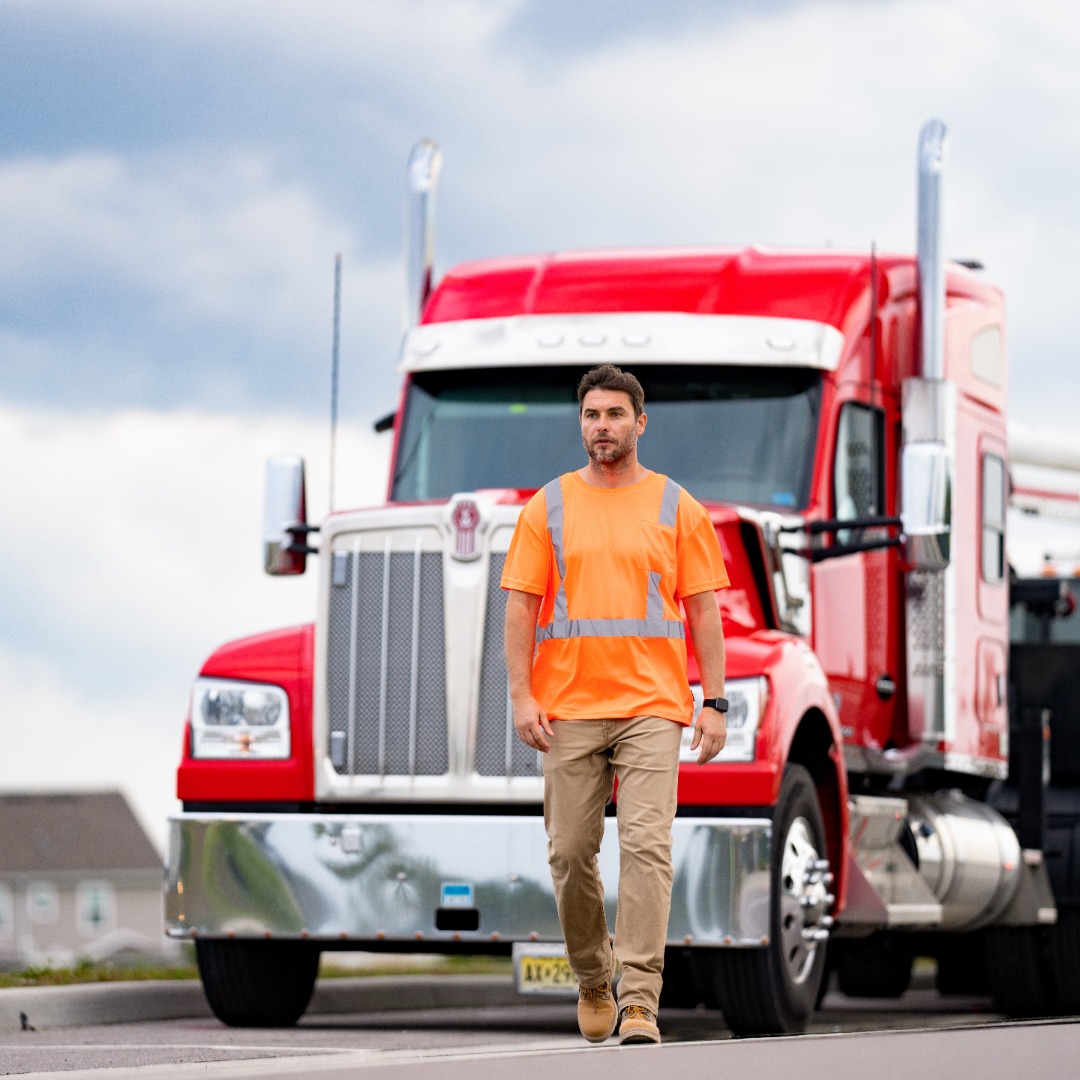 Man in an orange safety vest walking in front of a red semi-truck on a cloudy day.
