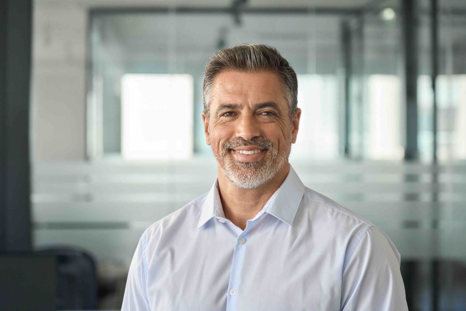 Smiling man with gray hair and beard in a white shirt standing in a modern office with glass walls.