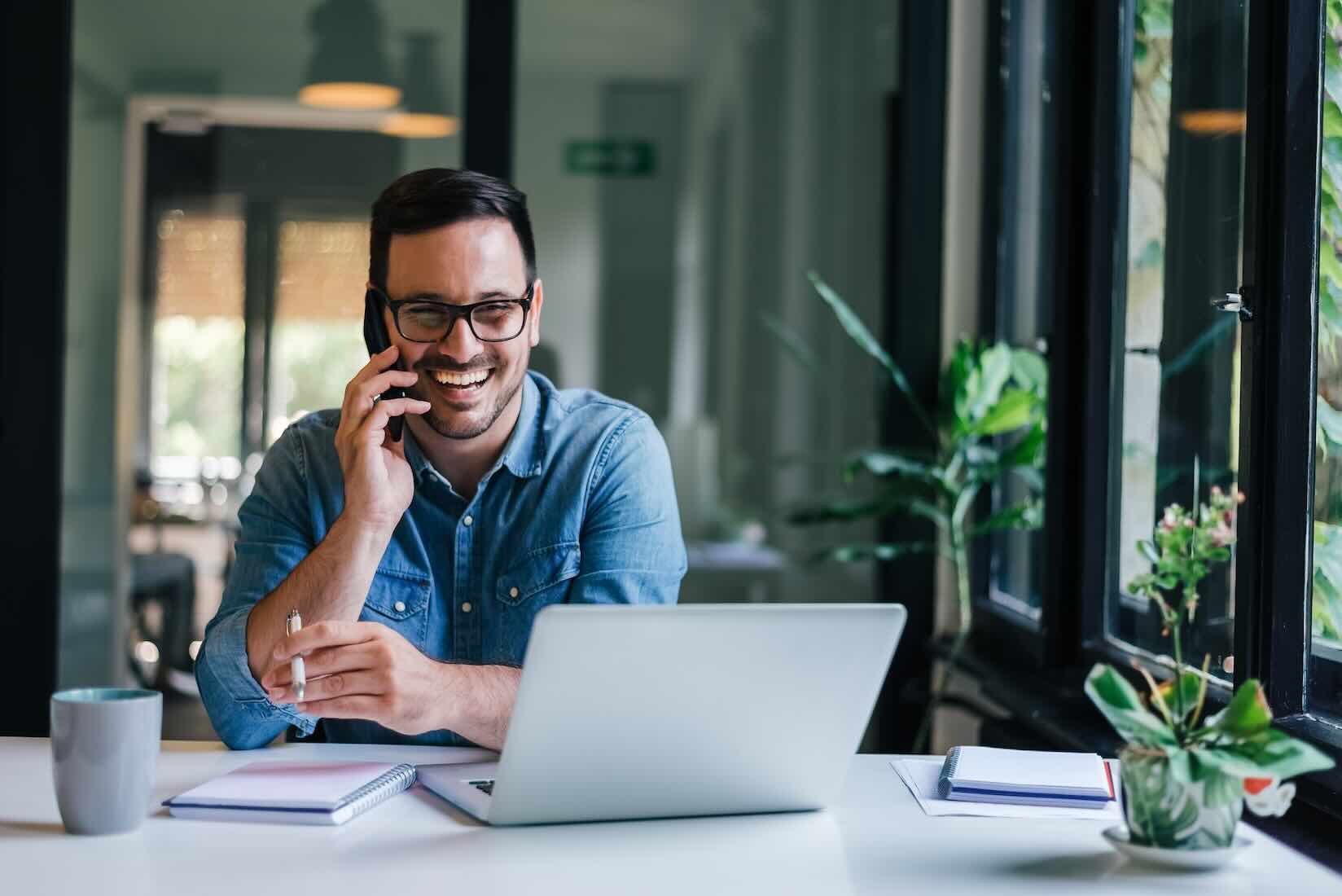 Smiling man in glasses talks on phone at a desk with a laptop, notebook, and coffee, near a window with plants.