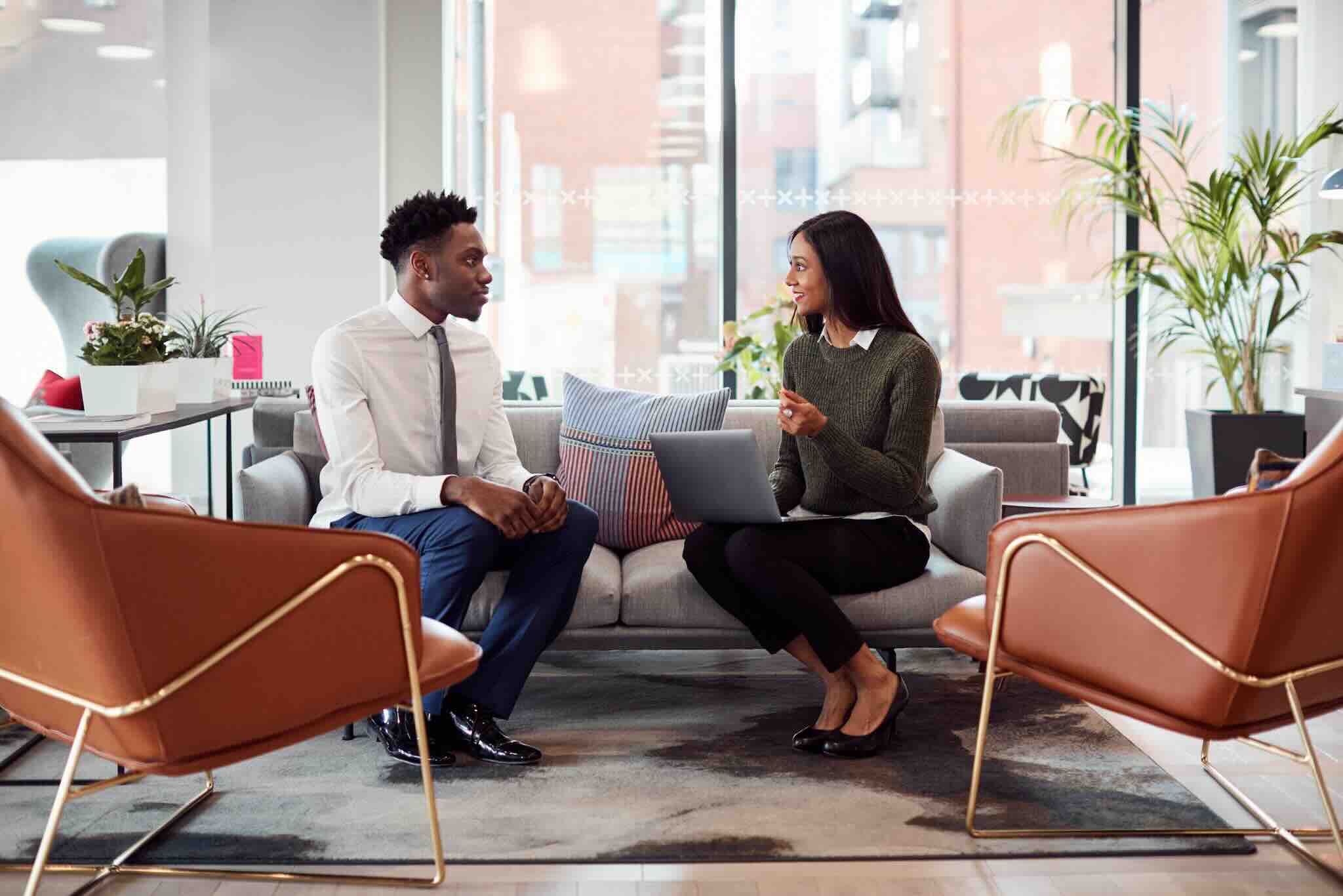 Two people sit on a couch in a modern office setting, discussing while one holds a laptop.