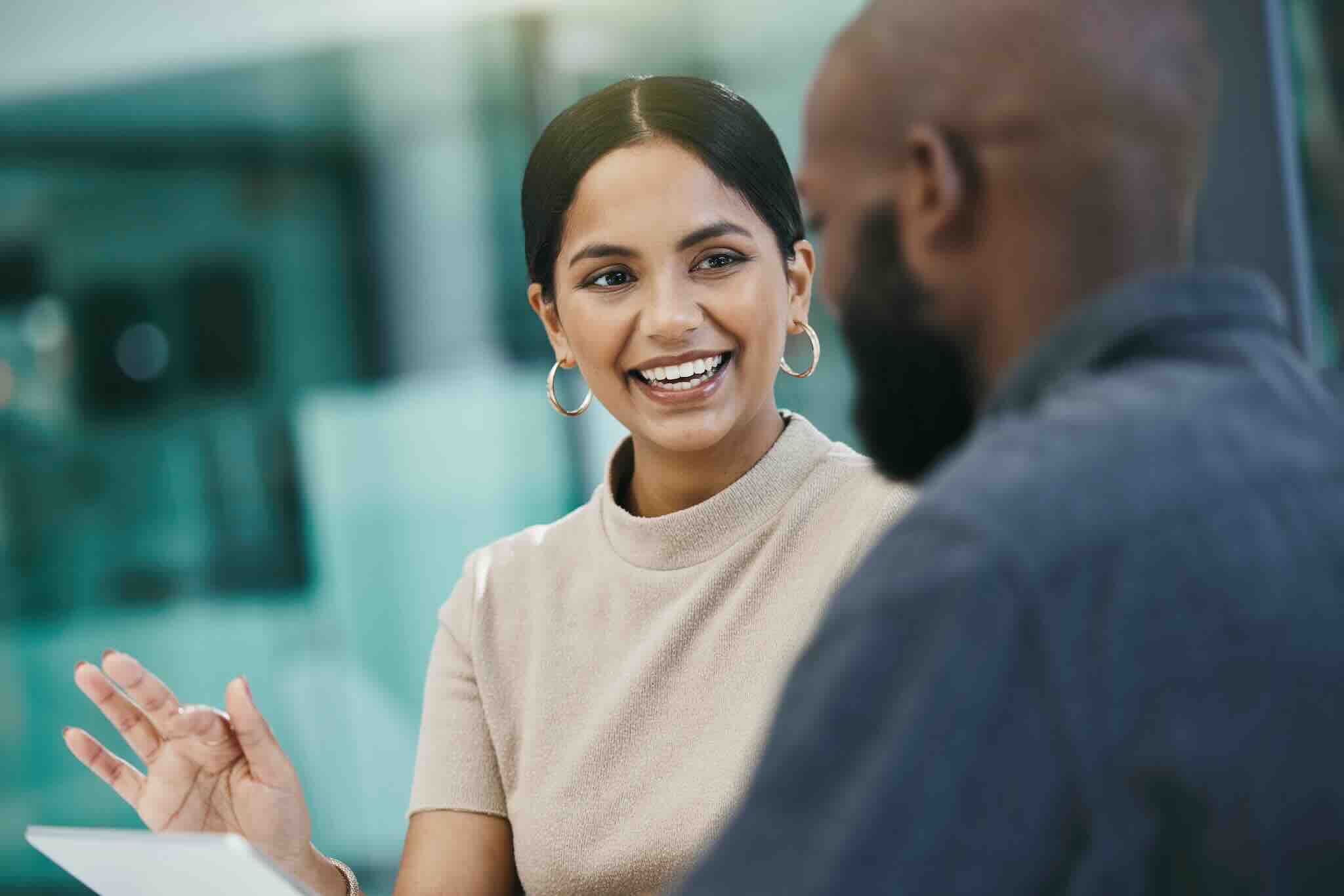 Smiling woman with hoop earrings engaging in conversation with a bald man in a business setting. She holds a tablet, appearing cheerful.