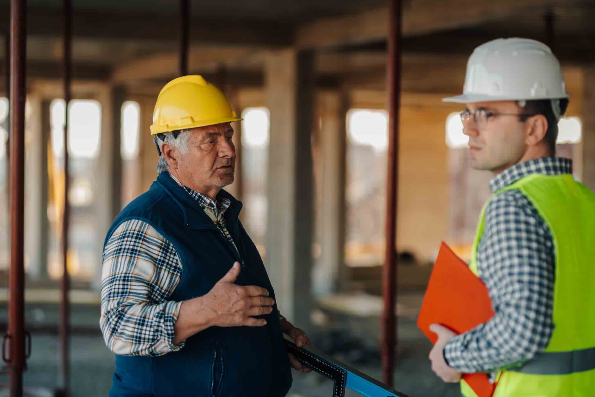 Two construction workers in hard hats and vests discussing at a job site. One holds a clipboard.