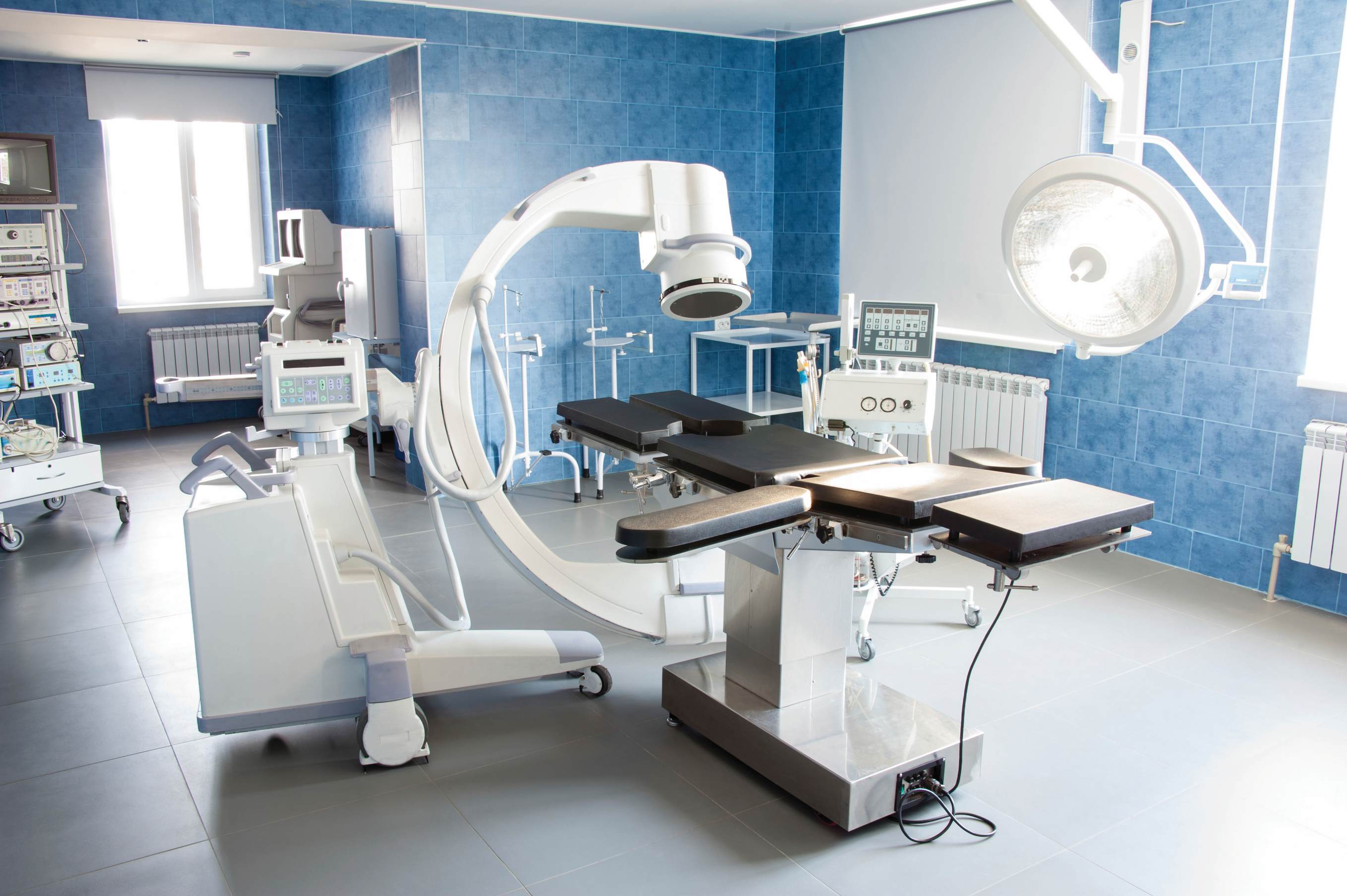 Modern hospital operating room with C-arm imaging, surgical table, and medical equipment against blue-tiled walls and bright lighting.