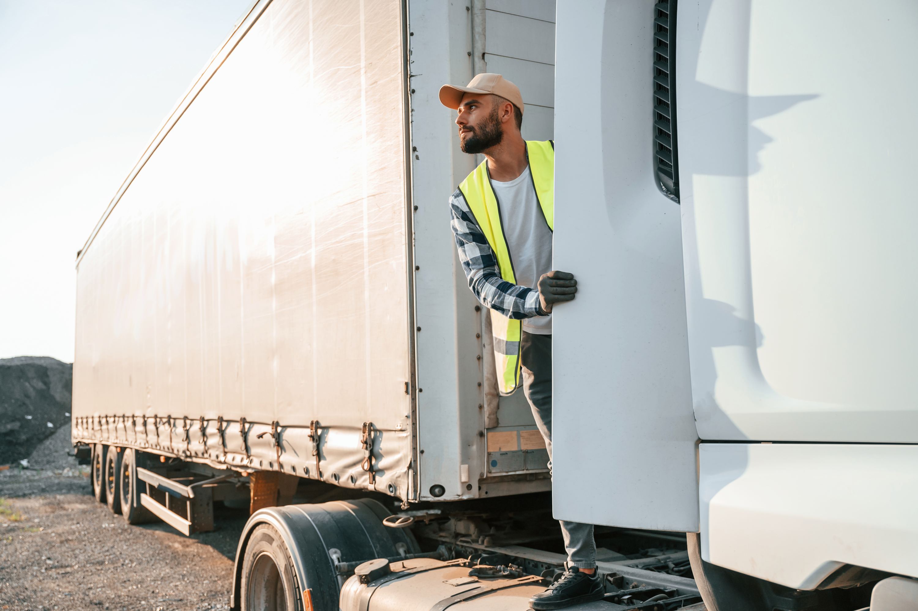 Worker in a high-visibility vest steps out of a truck cab, looking ahead, with a trailer in the background and morning light illuminating the scene.
