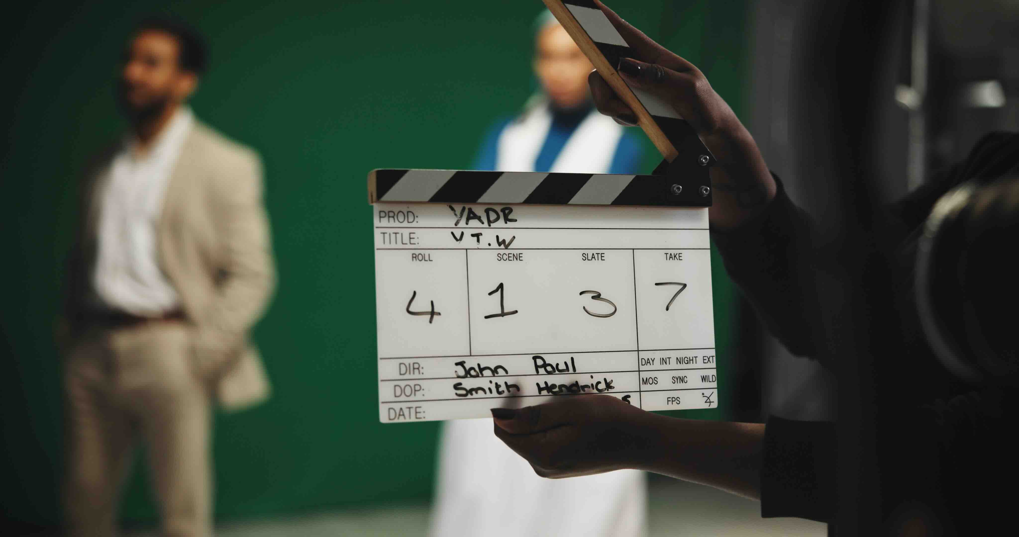 Film clapperboard held before a green screen, with blurred figures in the background.