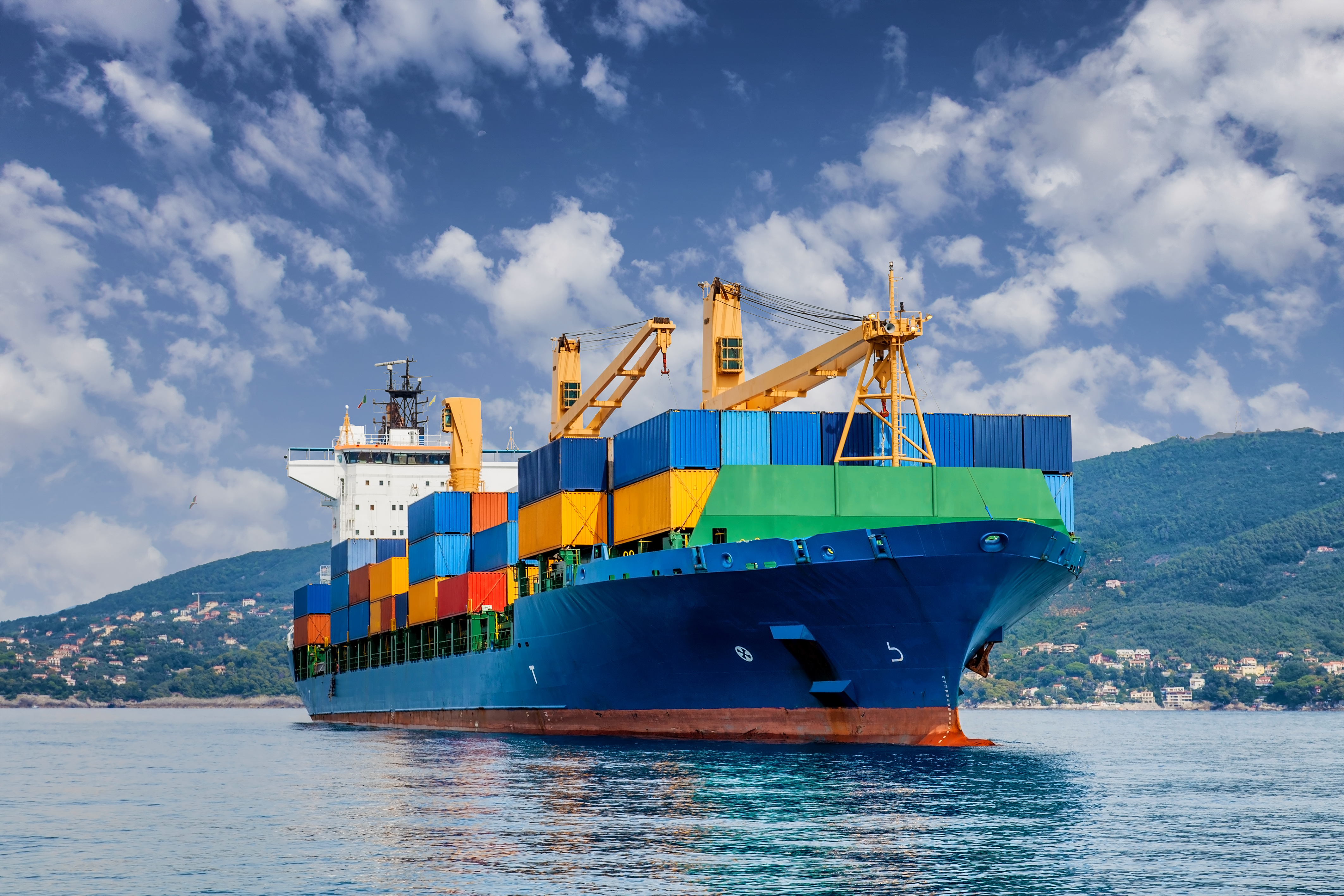 A large cargo ship with colorful containers sails under a partly cloudy sky, with a scenic hilly coastline in the background.