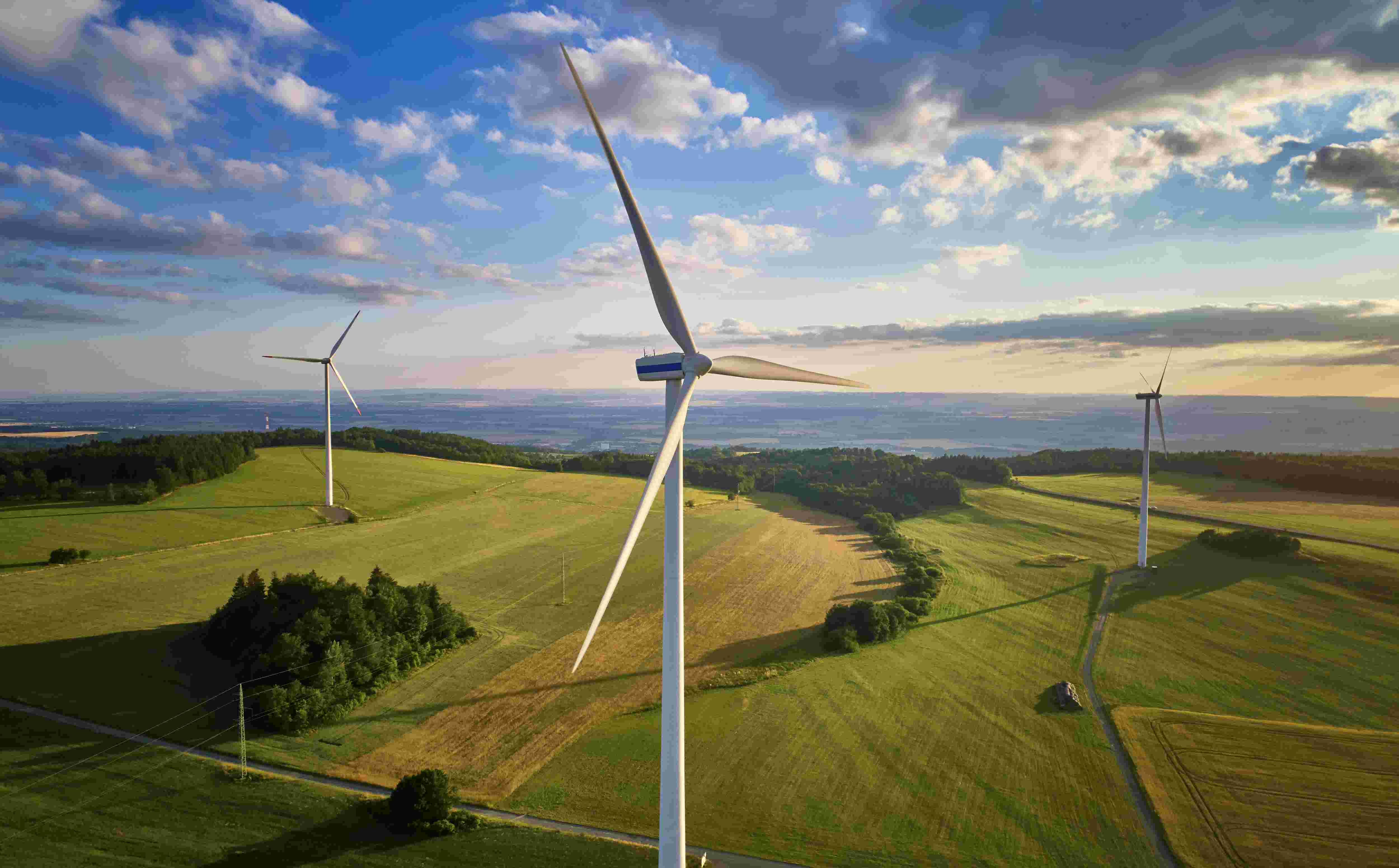 Three wind turbines on a green landscape under a partly cloudy sky, with fields stretching towards the horizon.