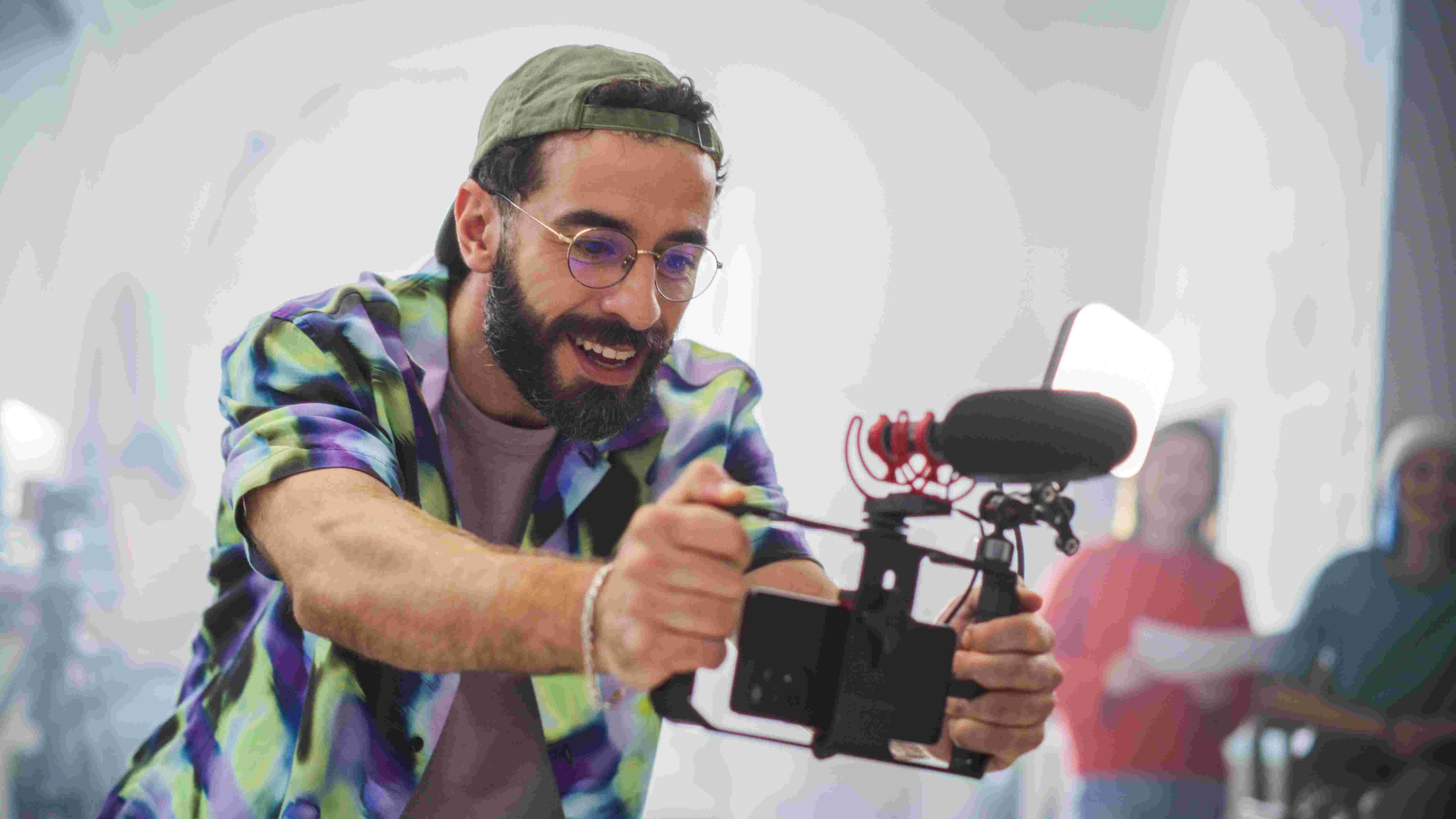Man in a colorful shirt and cap enthusiastically filming with a smartphone rig, featuring a microphone and light.