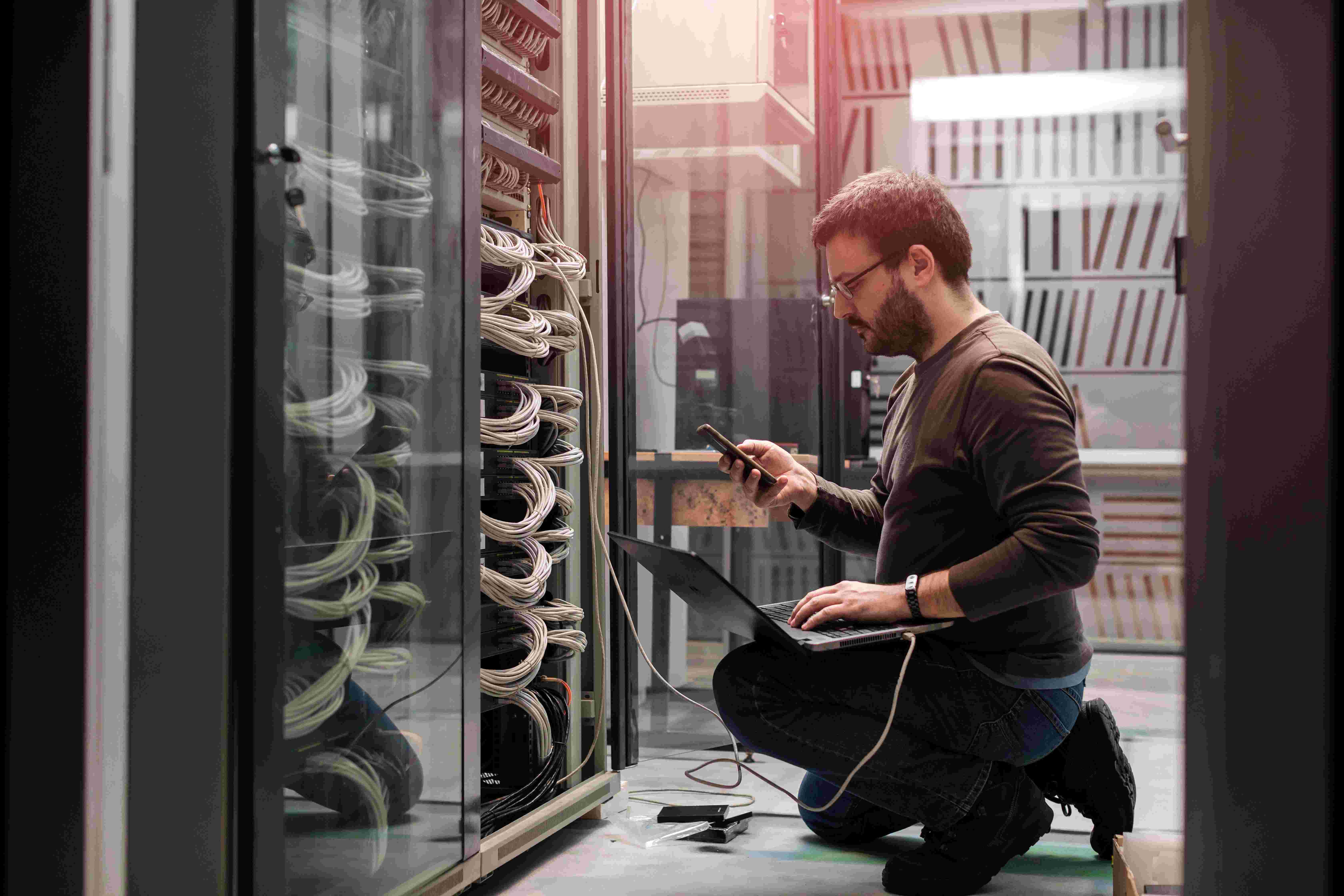 A person kneels in a server room, using a laptop and phone, surrounded by network cables.
