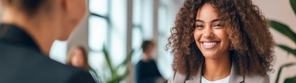 Two people in a bright office engaged in conversation. One person with curly hair is smiling warmly.