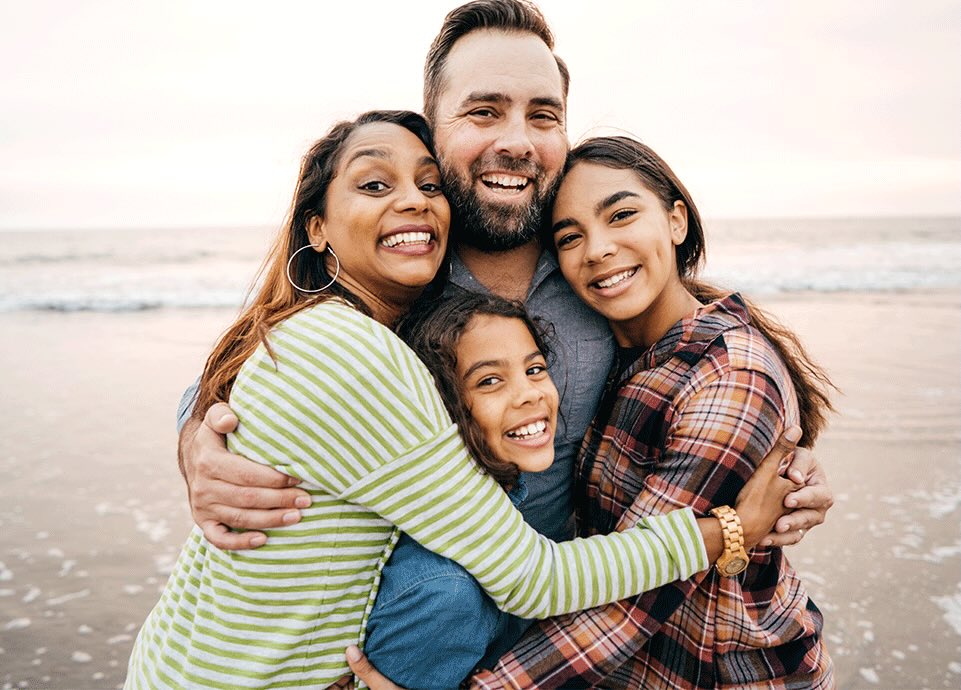 A joyful family of four embraces on a beach at sunset, smiling widely. The ocean waves gently approach the sandy shore in the background.