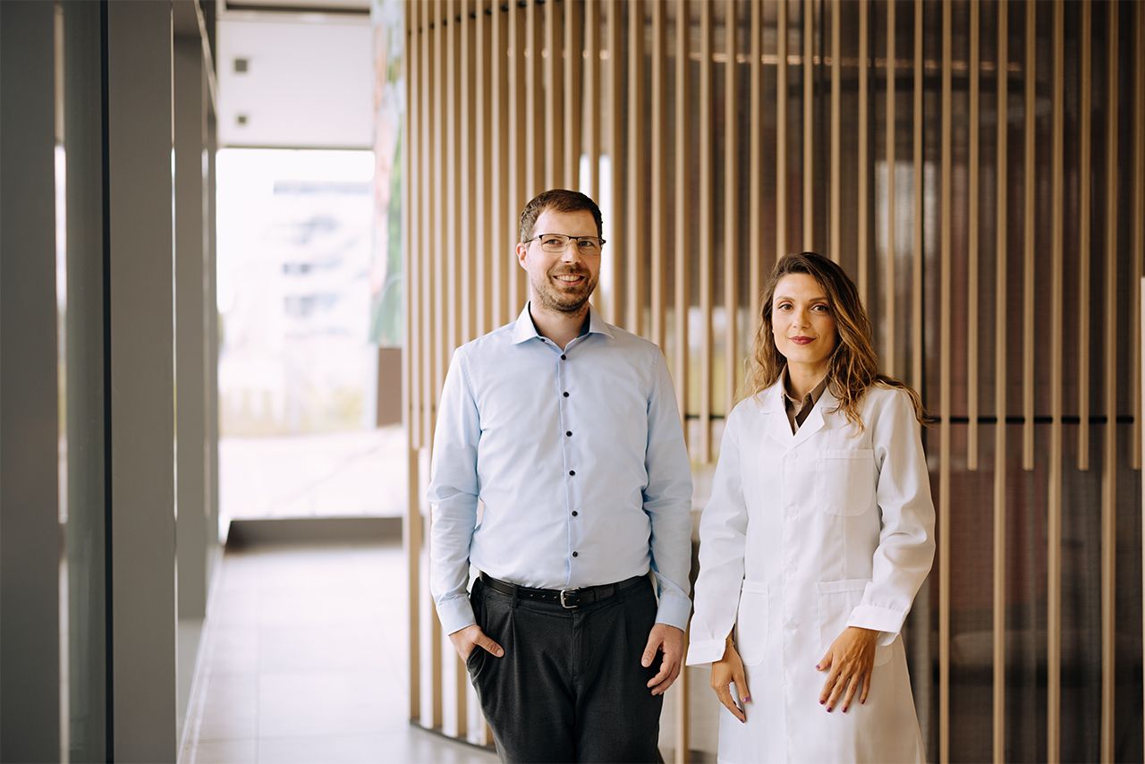 Ein Mann in einem hellblauen Hemd und eine Frau in einem weißen Laborkittel stehen nebeneinander in einem modernen Gebäude mit Holzverkleidung.